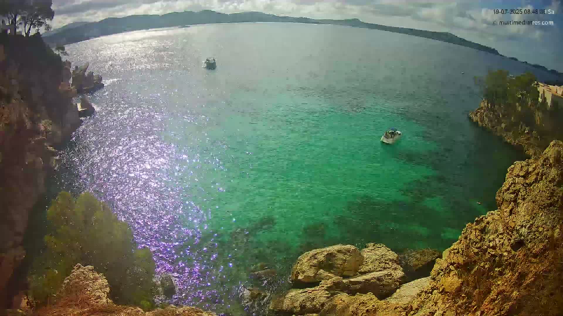 Sparkling, clear turquoise water laps against rocky shores under a partly cloudy sky, with a few small boats visible on the water.