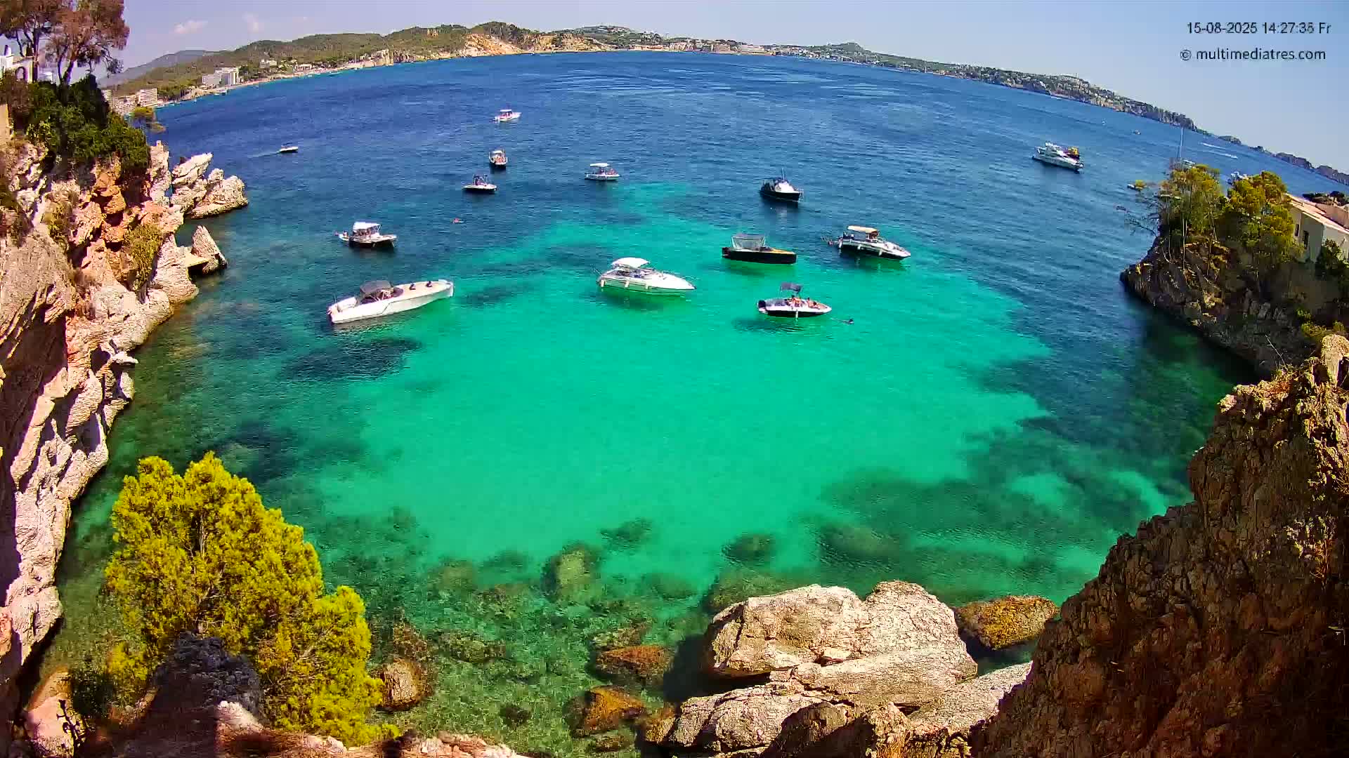A calm, sunny day shows clear turquoise water in a cove surrounded by rocky cliffs, with several boats anchored and people swimming.