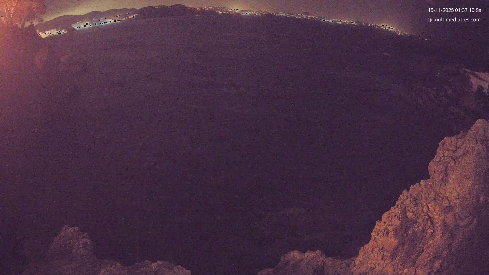 A wide-angle nighttime view shows a clear, dark sky over a rugged landscape with rocky formations in the foreground and distant lights of a town or city illuminating the horizon.