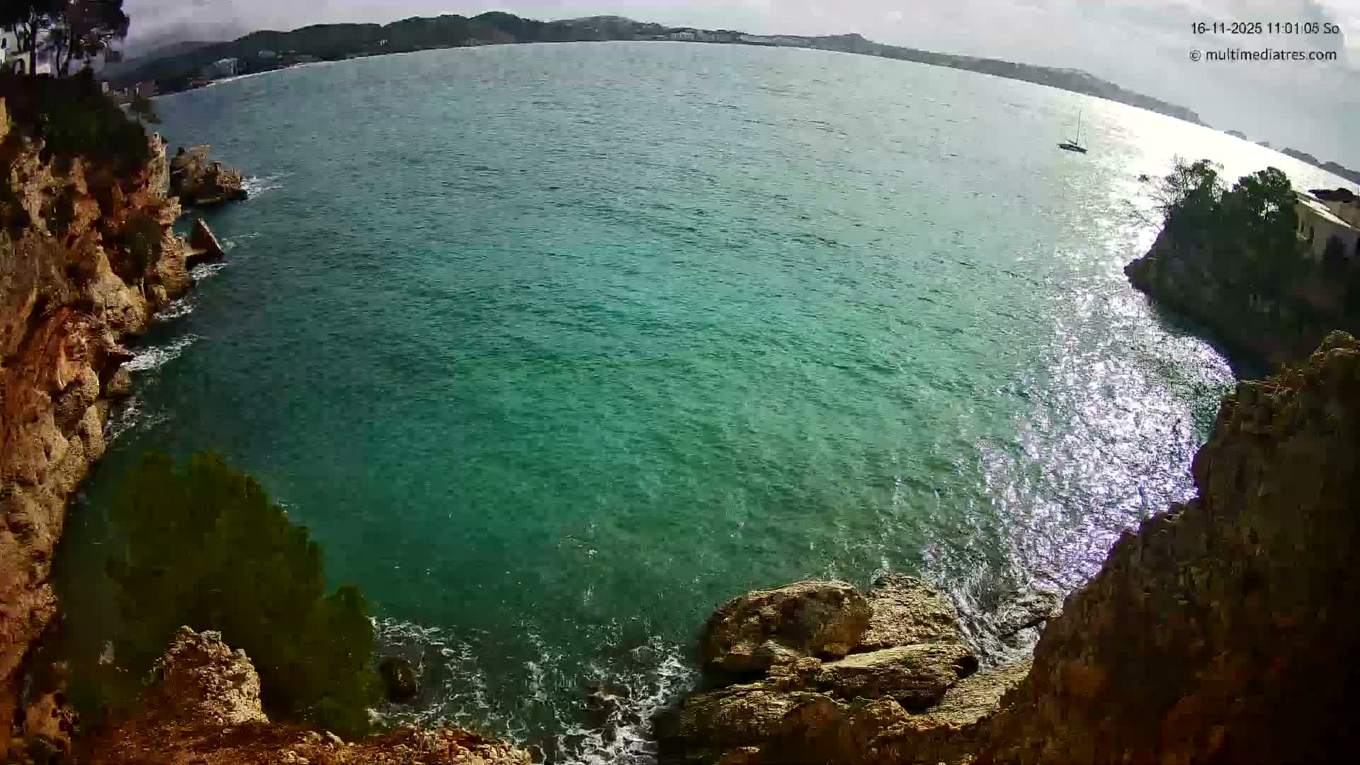 A wide-angle, elevated view showcases a bay with clear turquoise water, rugged rocky coastlines on either side, and a distant sailboat under a bright, overcast sky.