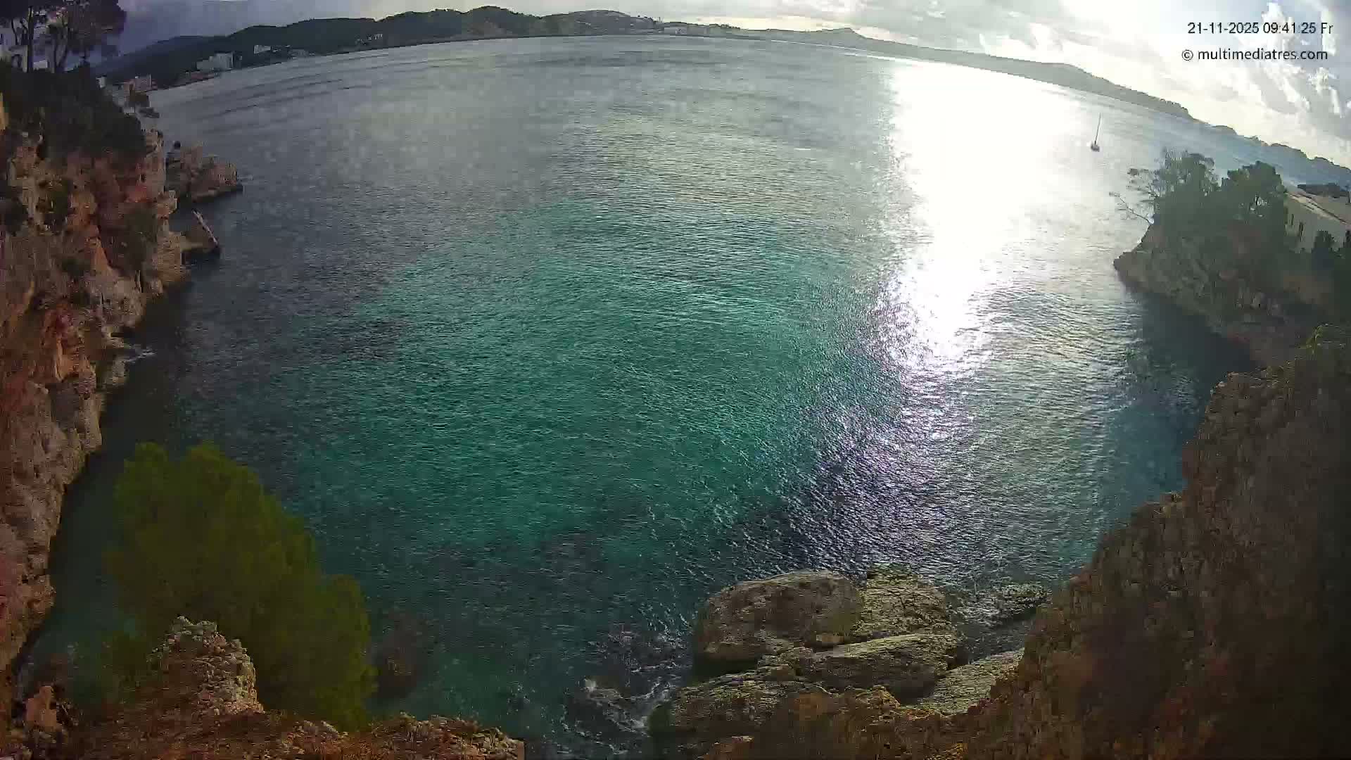 A bright, partially cloudy day illuminates a wide coastal bay with clear turquoise waters sparkling from the sun, flanked by rocky cliffs and distant hills with scattered buildings, featuring a lone sailboat on the calm sea.