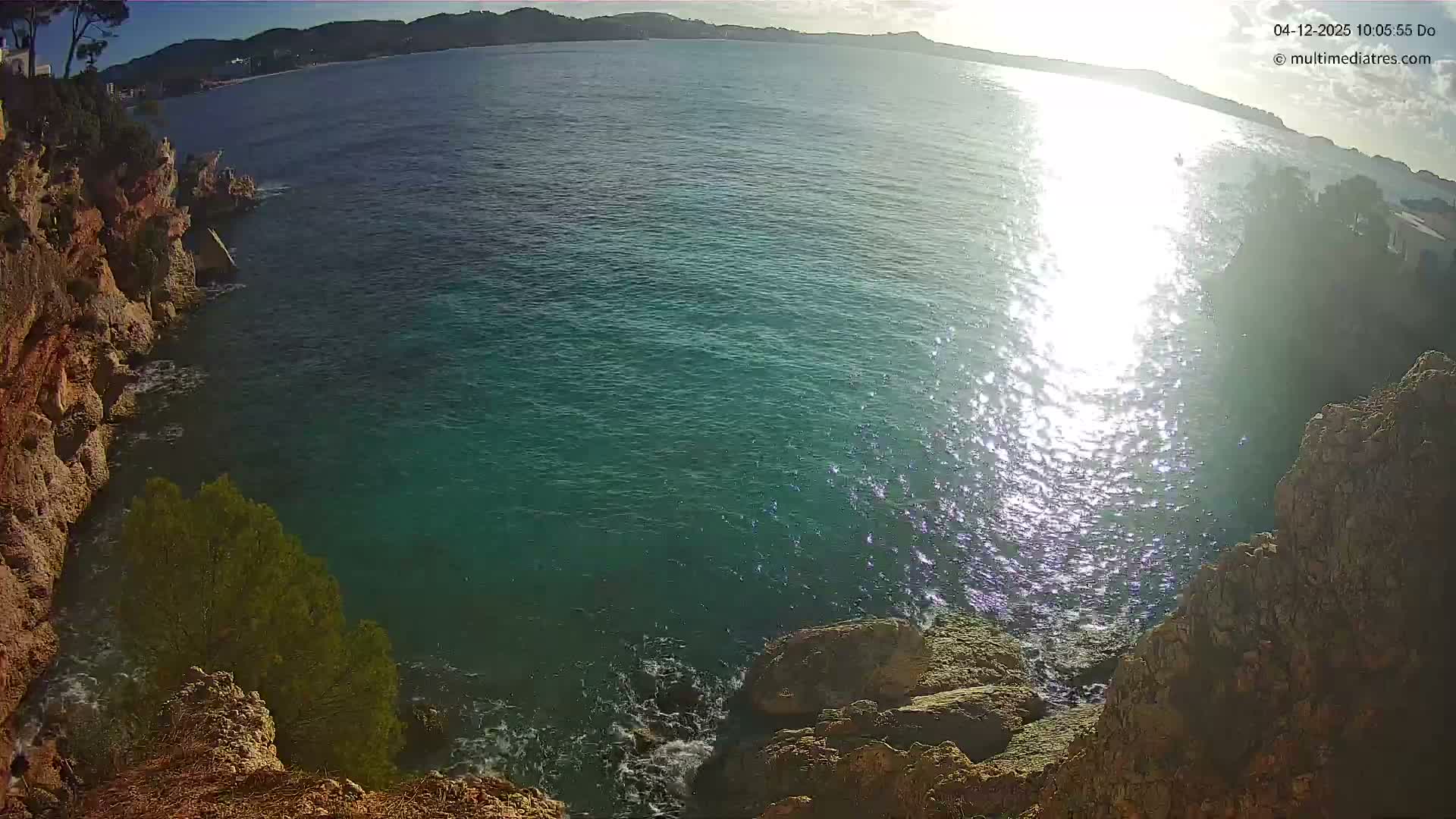 The image captures a sunny coastal scene with turquoise sea water reflecting bright sunlight, bordered by rugged cliffs on the left and foreground, and distant hills with a town visible under a clear sky.