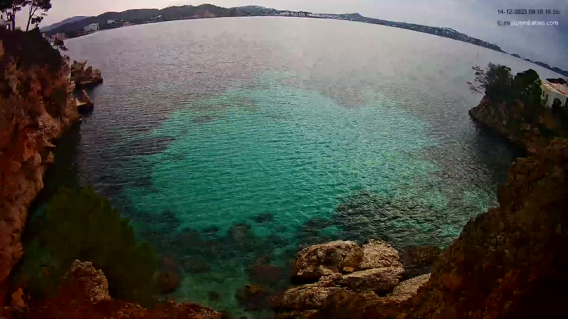 The image captures a sunny coastal scene with turquoise sea water reflecting bright sunlight, bordered by rugged cliffs on the left and foreground, and distant hills with a town visible under a clear sky.