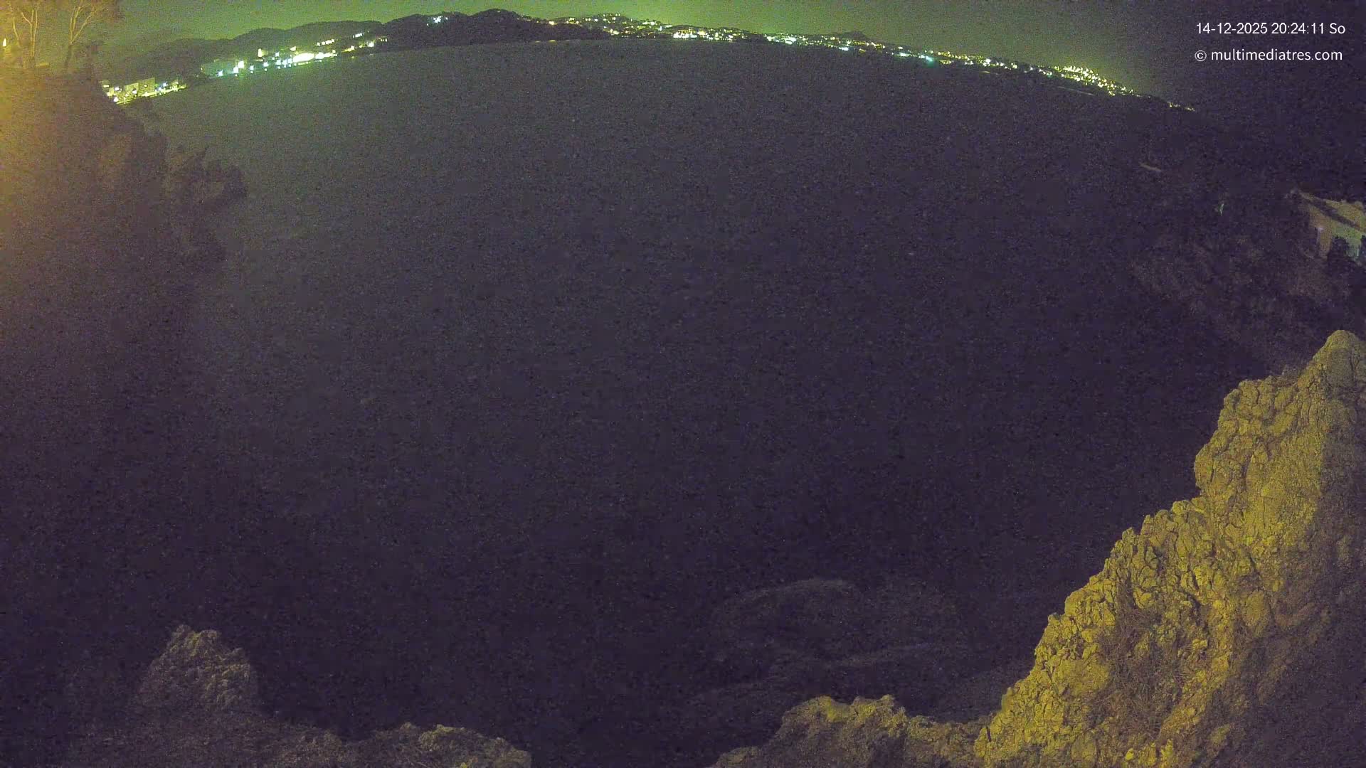 The image captures a sunny coastal scene with turquoise sea water reflecting bright sunlight, bordered by rugged cliffs on the left and foreground, and distant hills with a town visible under a clear sky.