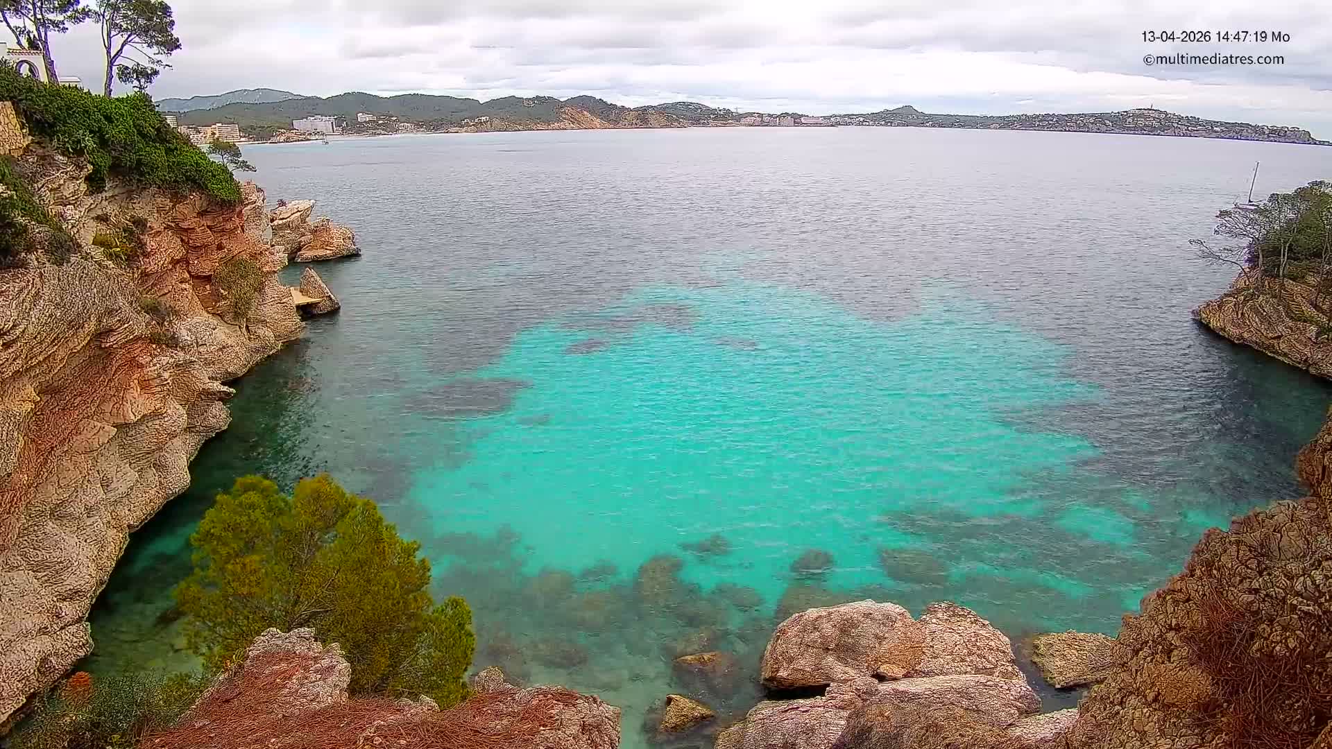 The image captures a sunny coastal scene with turquoise sea water reflecting bright sunlight, bordered by rugged cliffs on the left and foreground, and distant hills with a town visible under a clear sky.