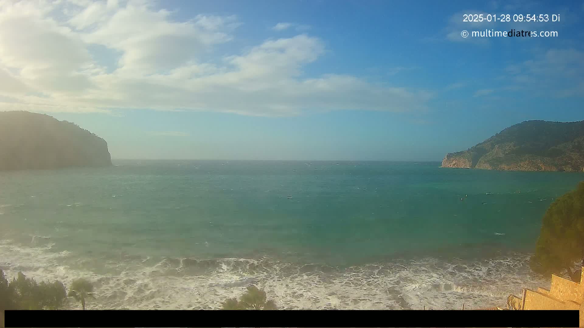 A partly cloudy day shows a wide expanse of choppy teal ocean water between two dark-green hills, with some waves crashing on the shore in the foreground.