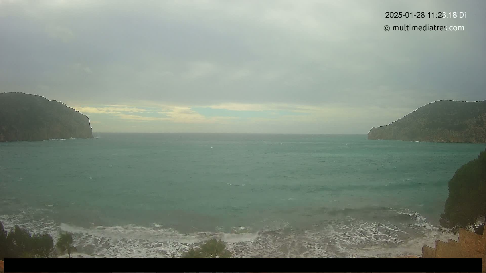 A choppy teal sea, under a cloudy sky, is nestled between two dark, rocky hillsides, with waves breaking on a nearby shore.