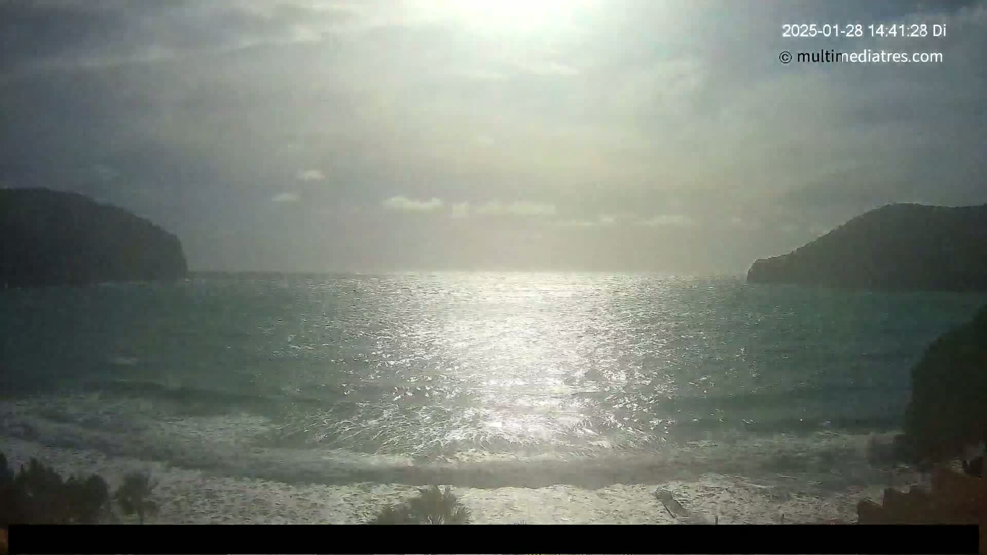 A bright, somewhat hazy sun shines over a wavy sea between two dark, distant hills.
