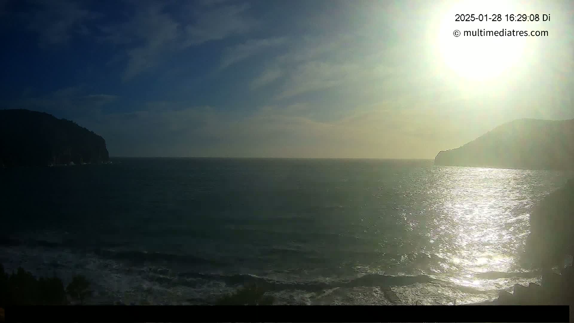 A bright sun shines over a calm sea with small waves, between two dark hills or mountains under a partly cloudy sky.