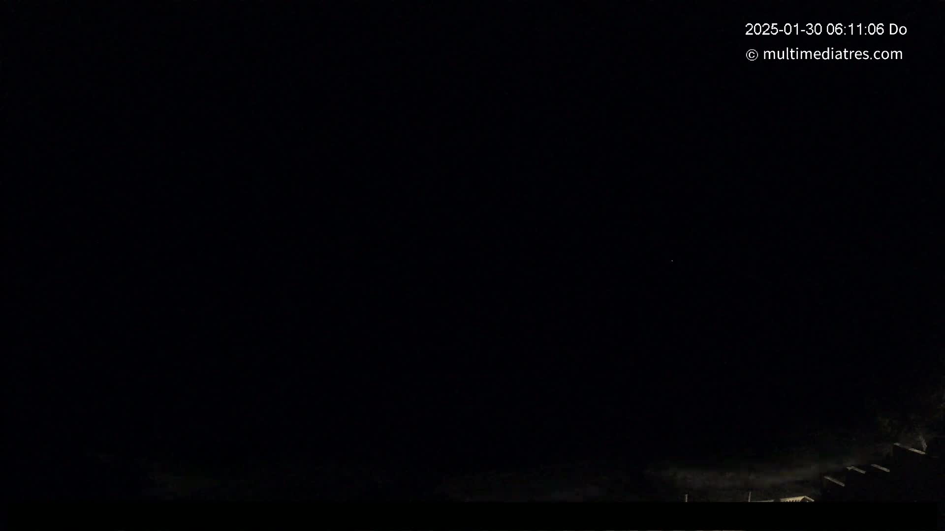 A nighttime, dark image shows faintly lit structures at the bottom edge of the frame under a mostly clear, dark sky.