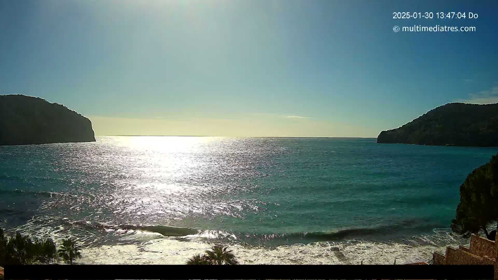 A sunny day reveals a tranquil bay with clear turquoise water lapping a sandy shore, framed by two dark, rocky hills.