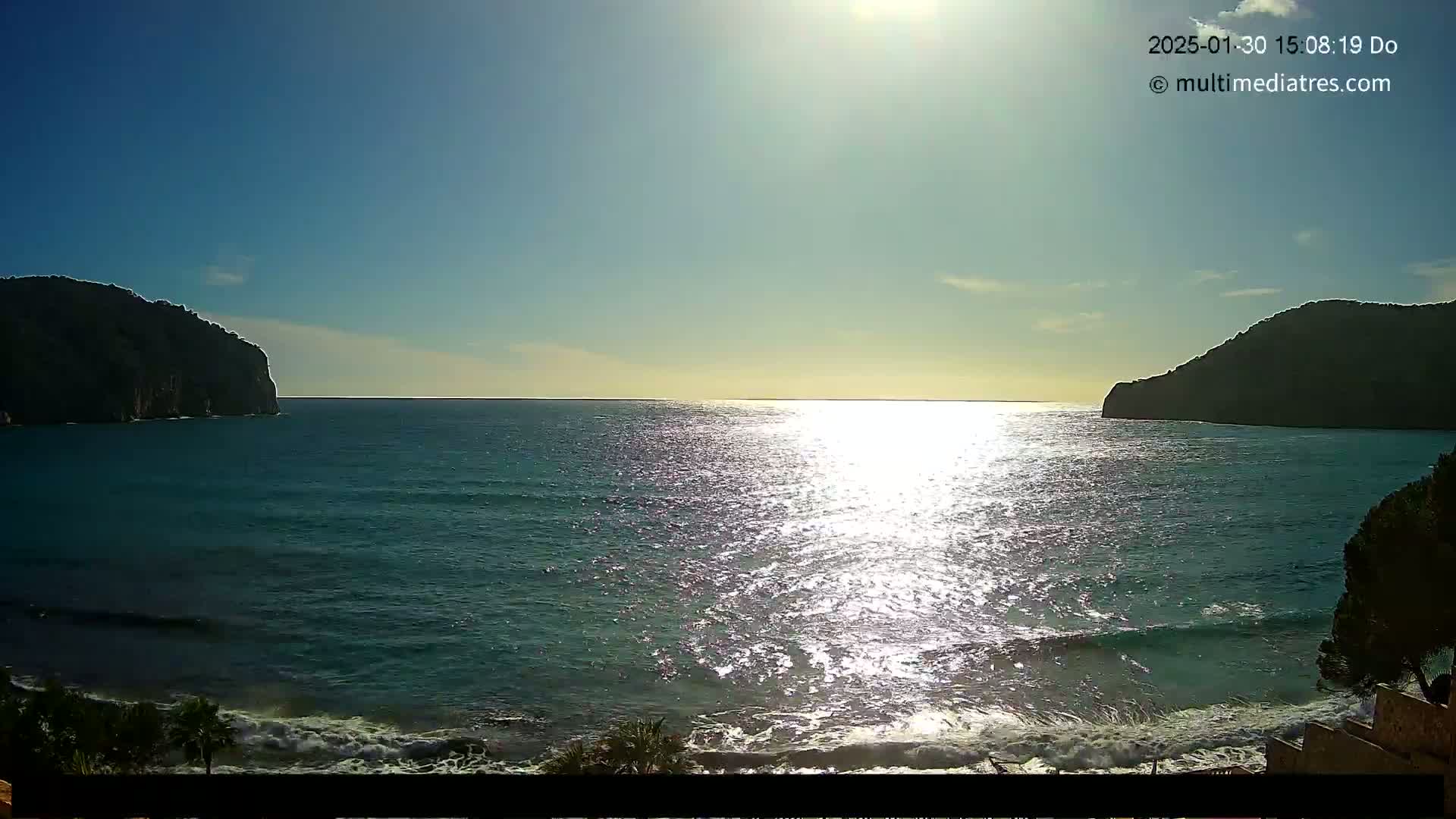 A bright, sunny day shows a calm sea with small waves lapping a shore between two dark, rocky headlands.