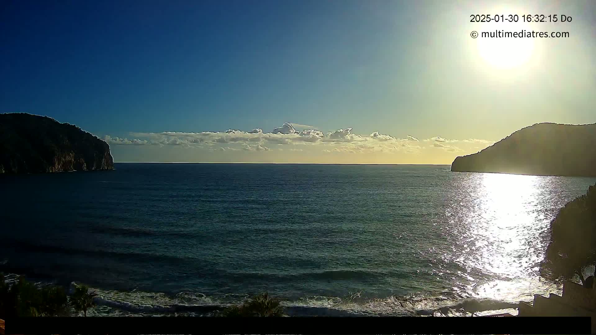 A calm sea with a partly cloudy sky and bright sunlight reflecting on the water, framed by dark hills on either side.