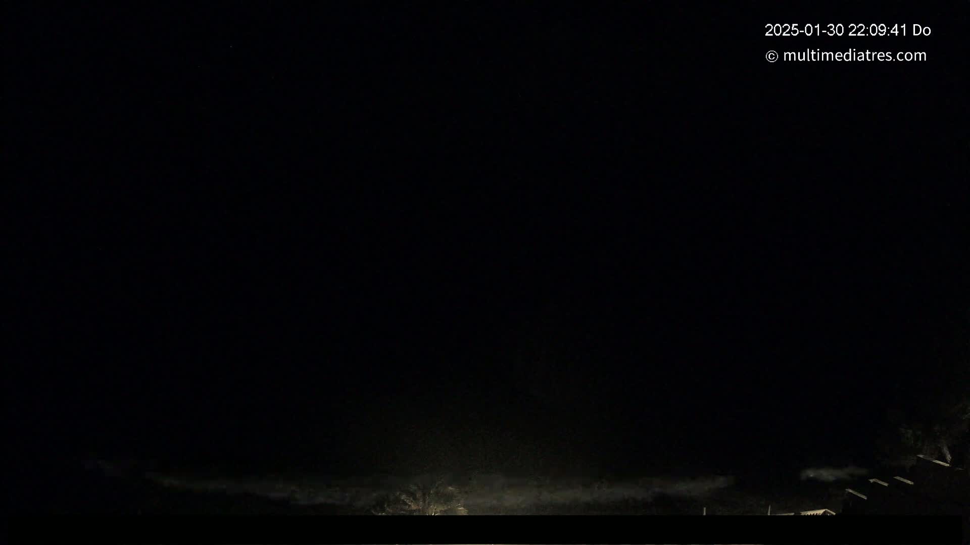 A nighttime, dark outdoor scene shows a low-lying fog or mist over a flat landscape, with dimly lit structures visible in the lower right corner.