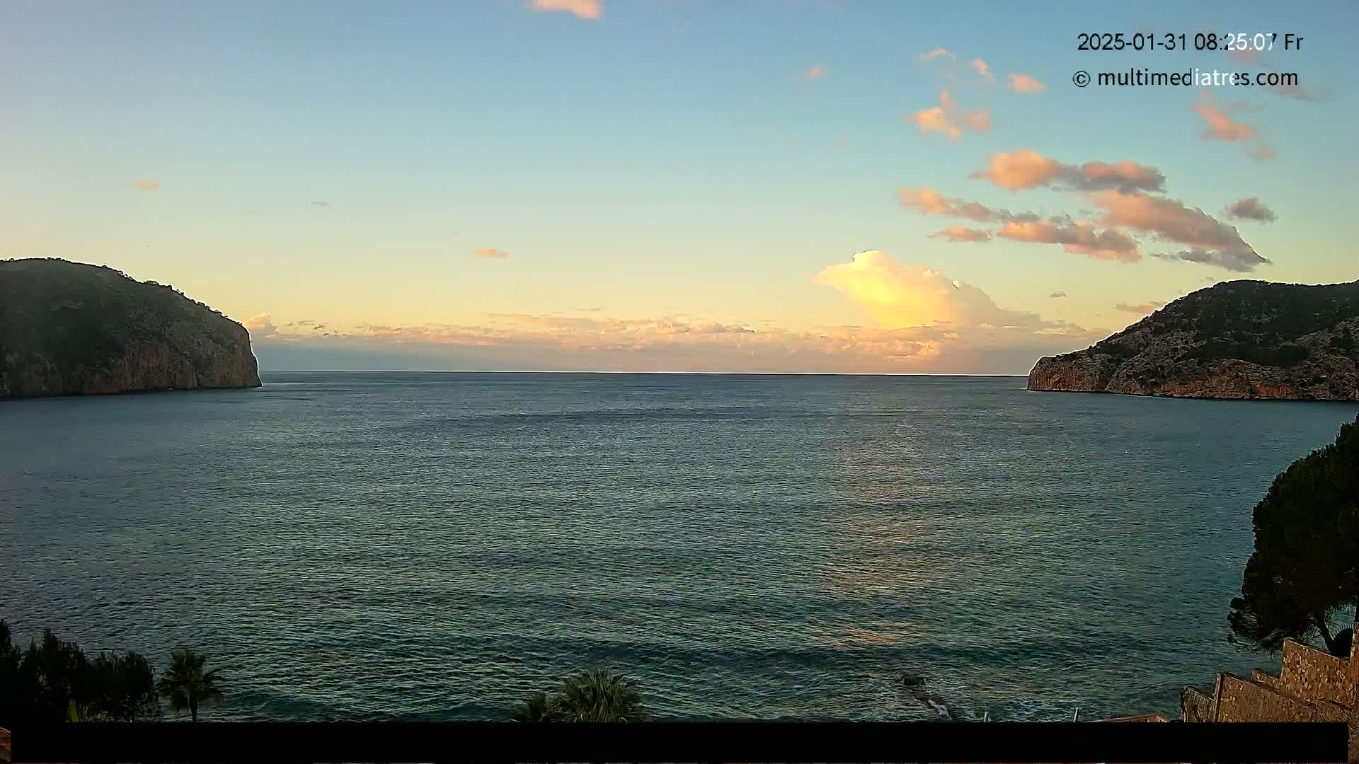 A calm sea at sunrise is nestled between two dark cliffs under a partly cloudy sky.
