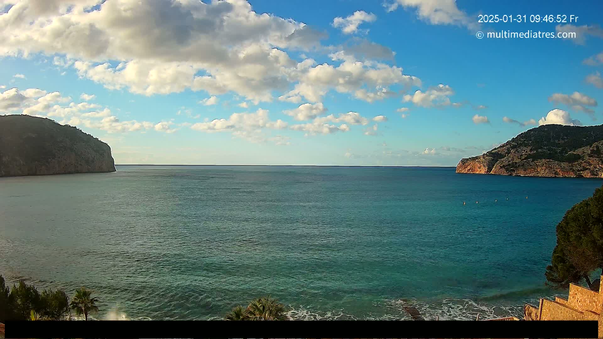 A partly cloudy day shows a tranquil teal sea nestled between two rocky headlands, with a small section of beach and vegetation visible in the foreground.