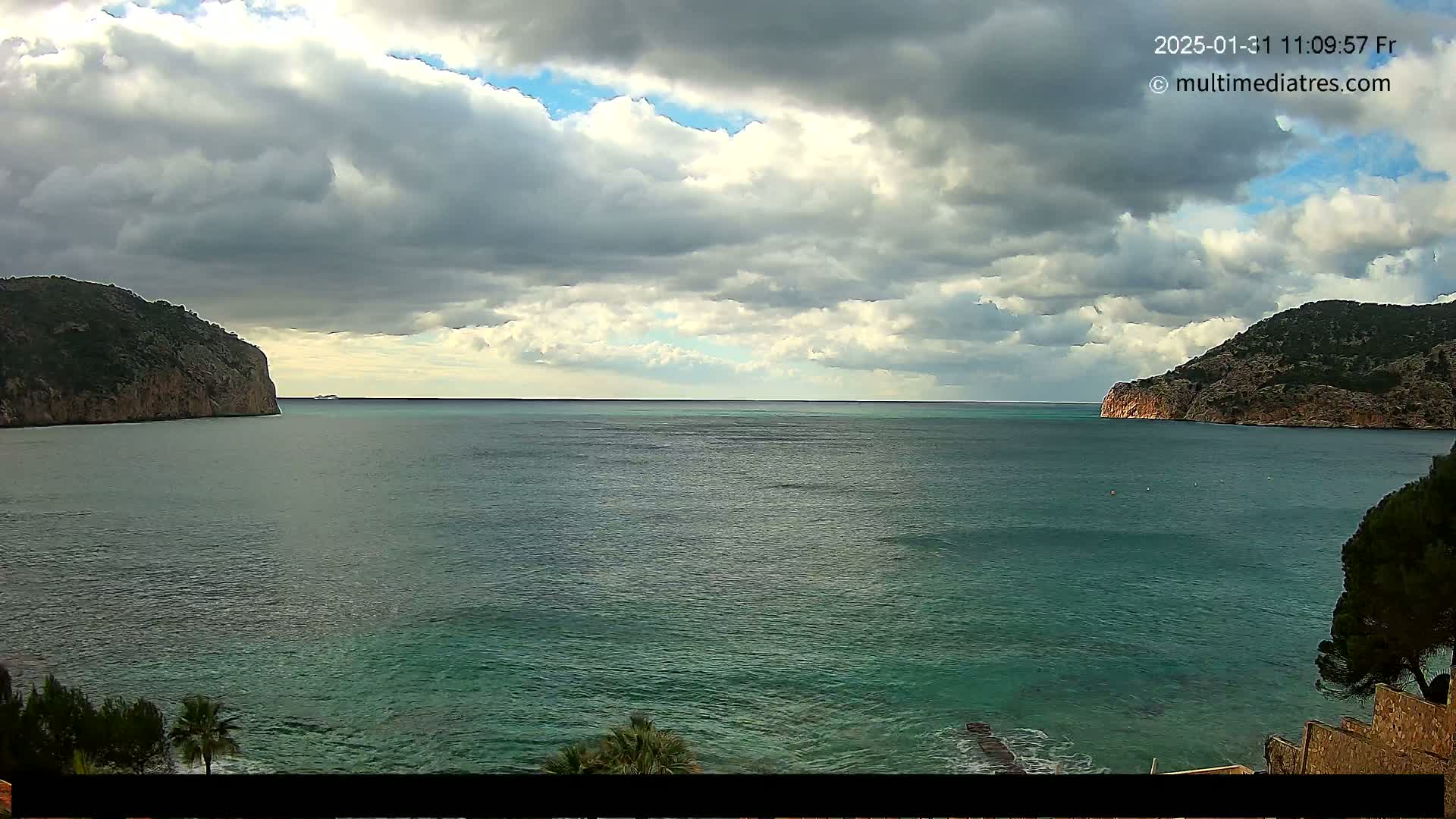 A partly cloudy sky overlooks a calm teal sea, bordered by two rocky headlands.
