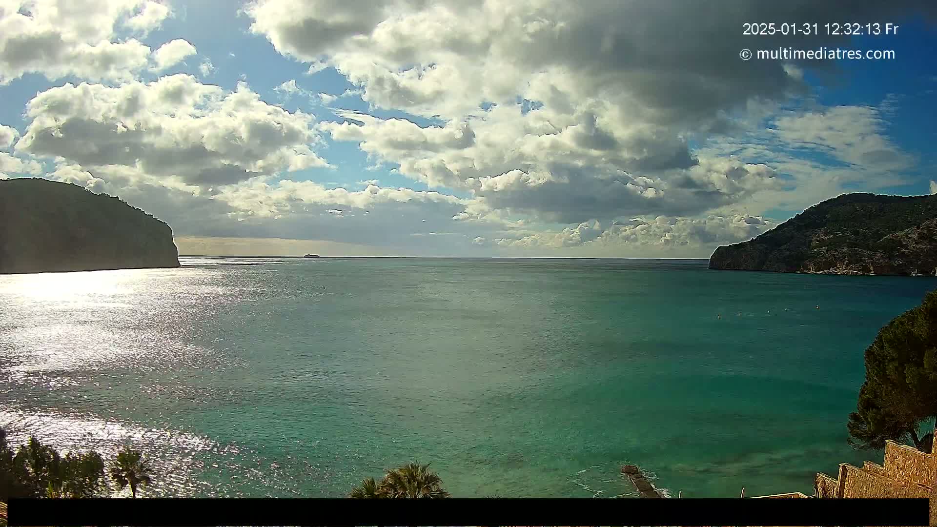 A partly cloudy sky overlooks a calm, teal ocean bay nestled between two dark green hills.