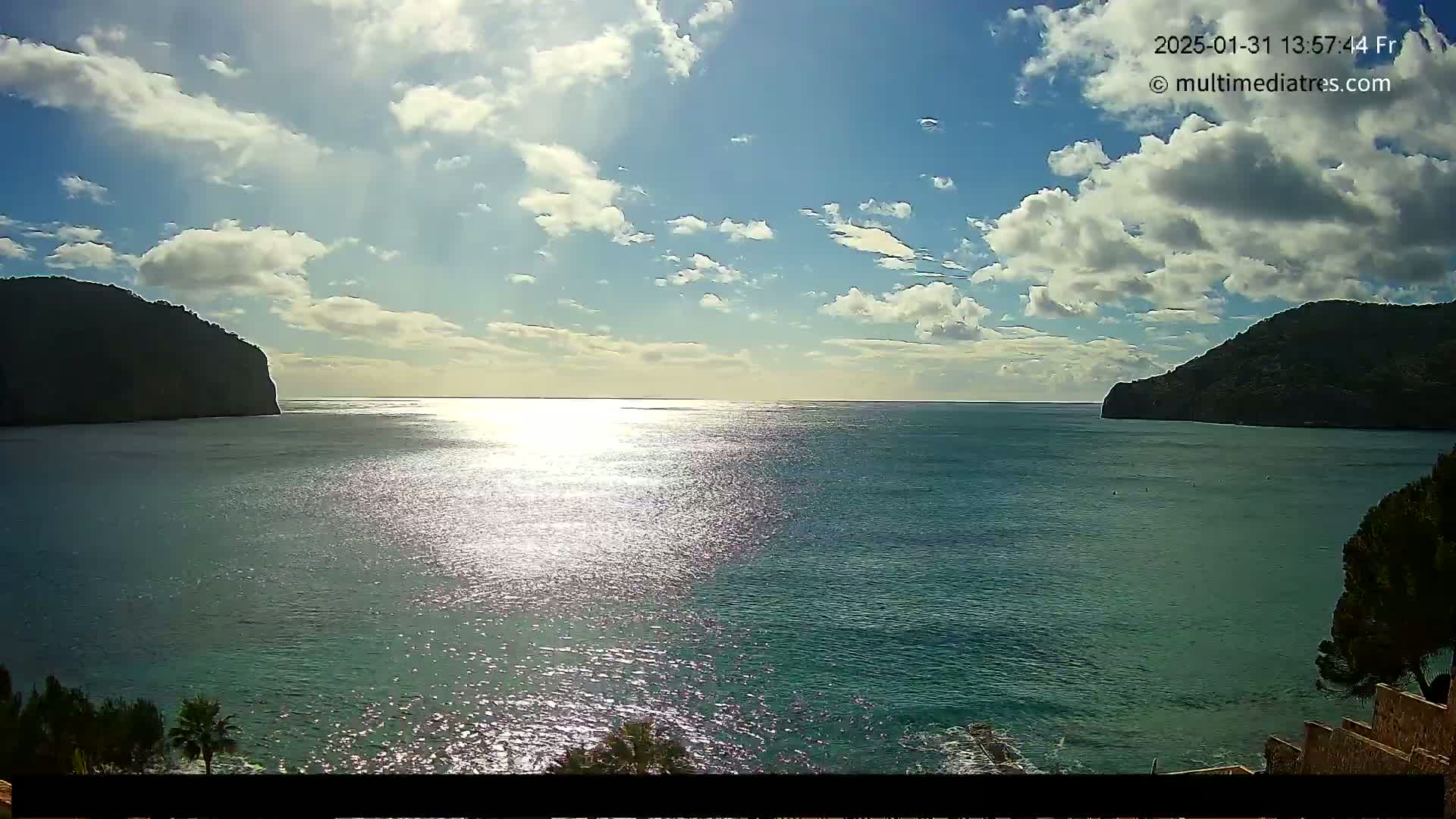A partly sunny day shows a calm sea separating two dark, rocky hillsides under a mostly cloudy sky.