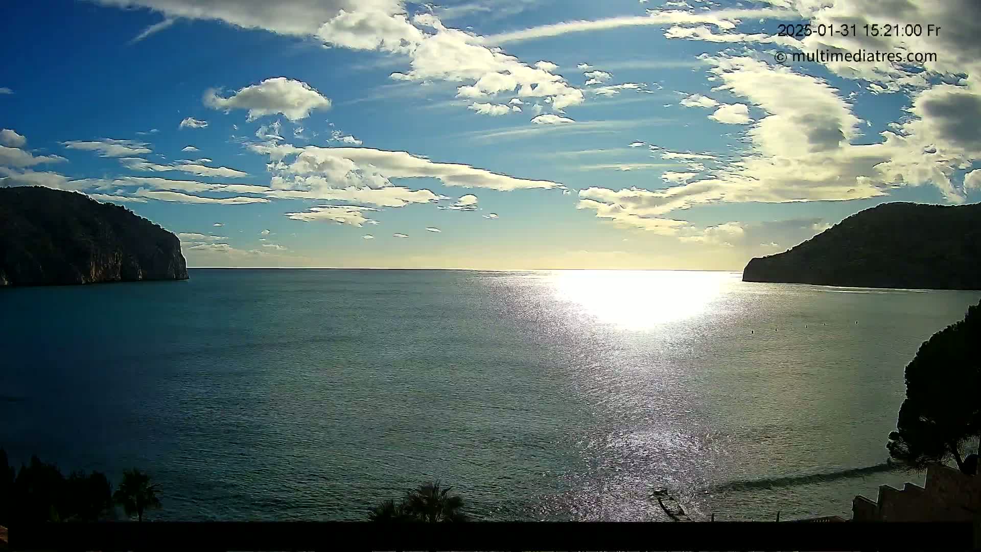 A calm sea reflects sunlight under a partly cloudy sky, with dark hills or mountains on either side of the bay.