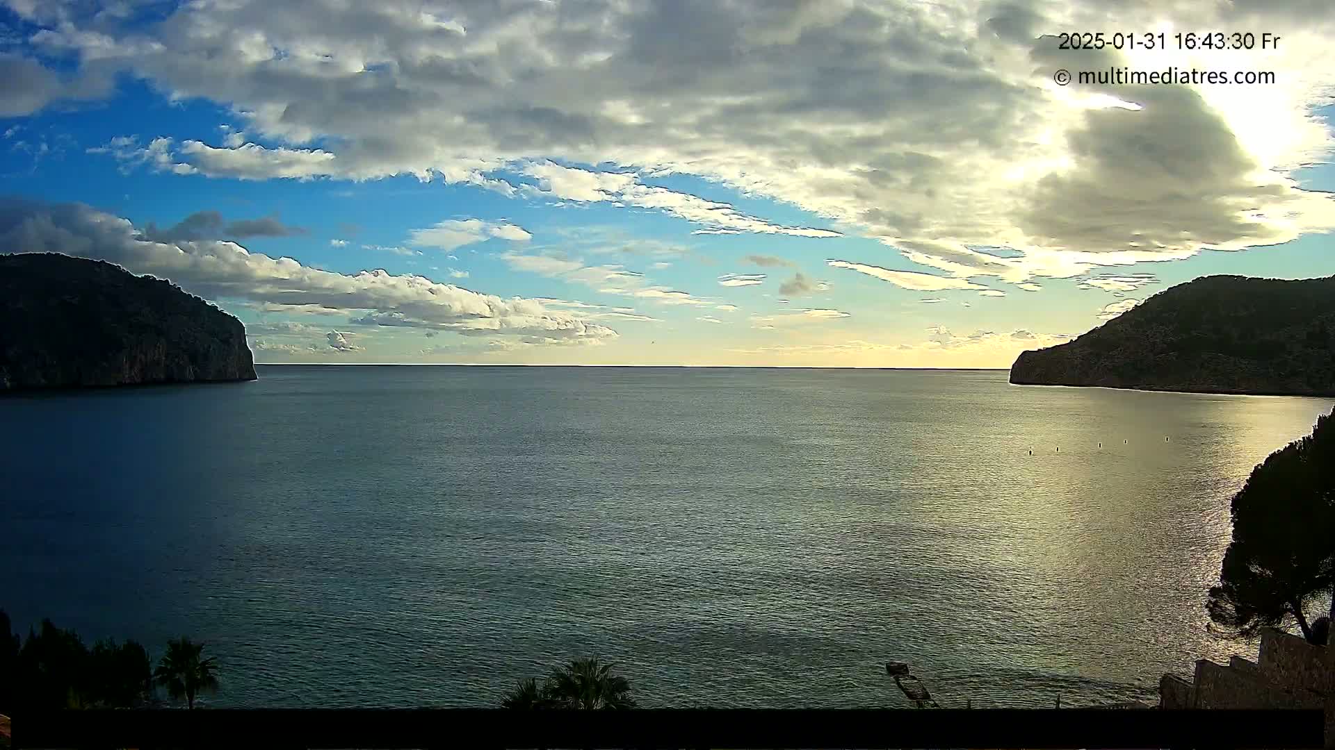 A partly cloudy sky overlooks a calm sea between two dark, rocky headlands.
