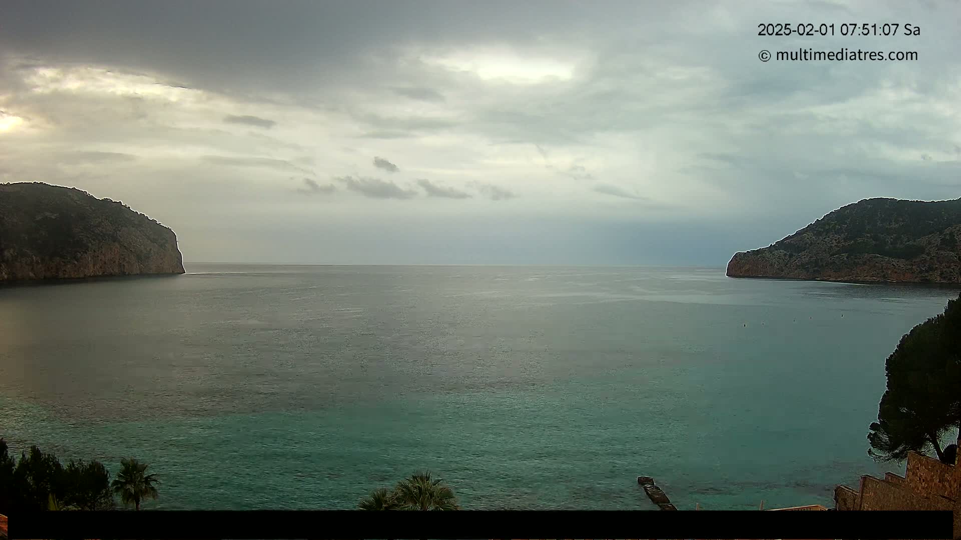 A calm, overcast day shows a bay of teal water between two rocky, dark-green hillsides.