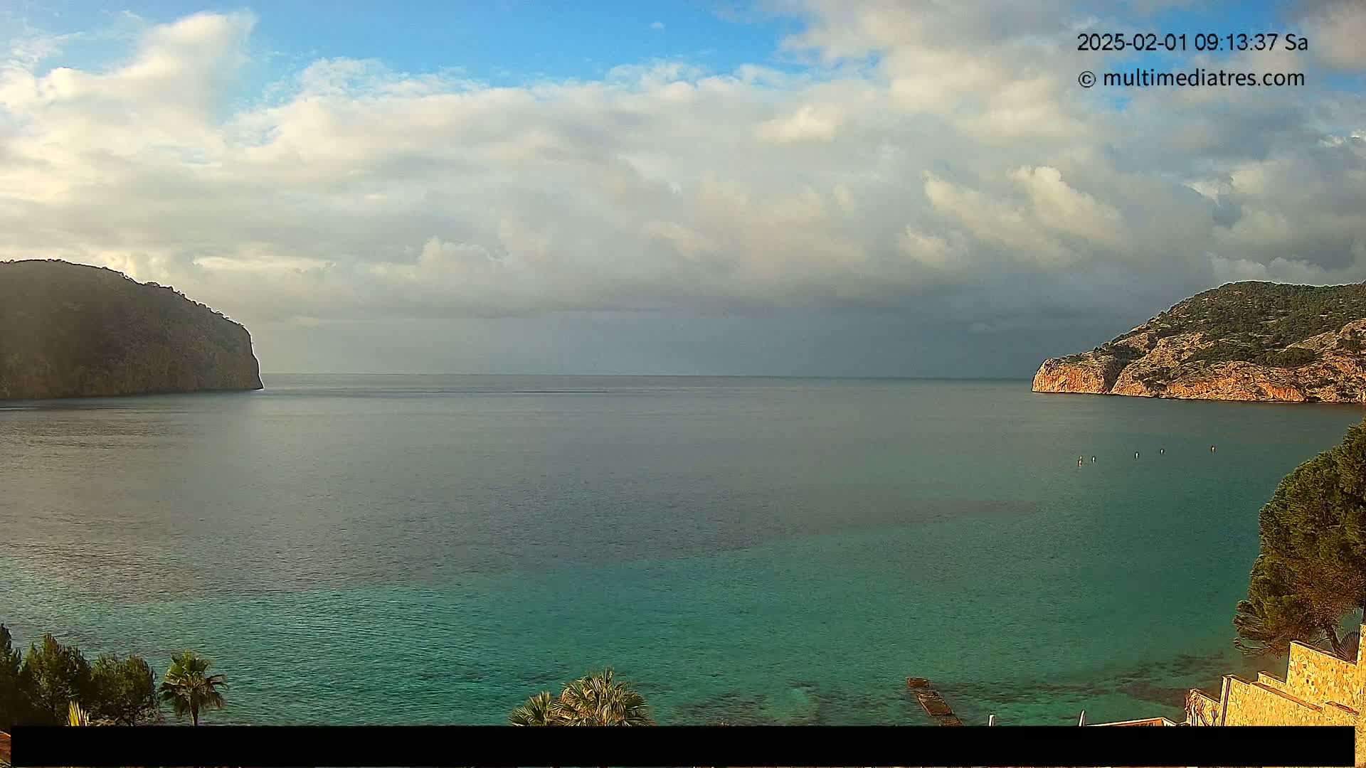 A partly cloudy day reveals a tranquil bay with calm, teal-colored water nestled between two rocky headlands.