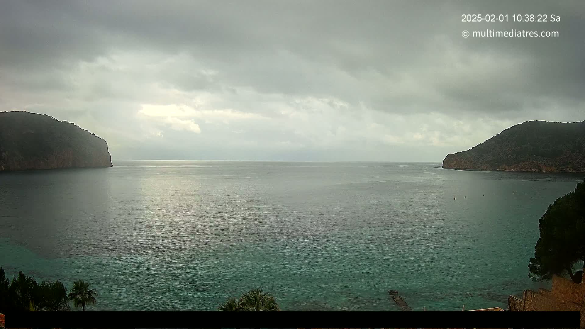 A body of calm, teal water is nestled between two dark, rocky hills under an overcast sky.