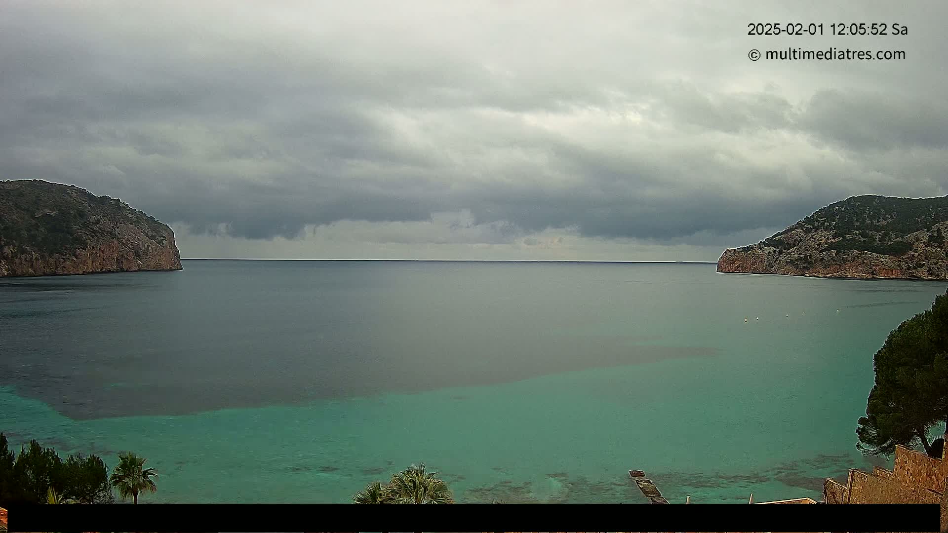 A calm, teal-colored bay is nestled between two rocky hills under a cloudy sky.
