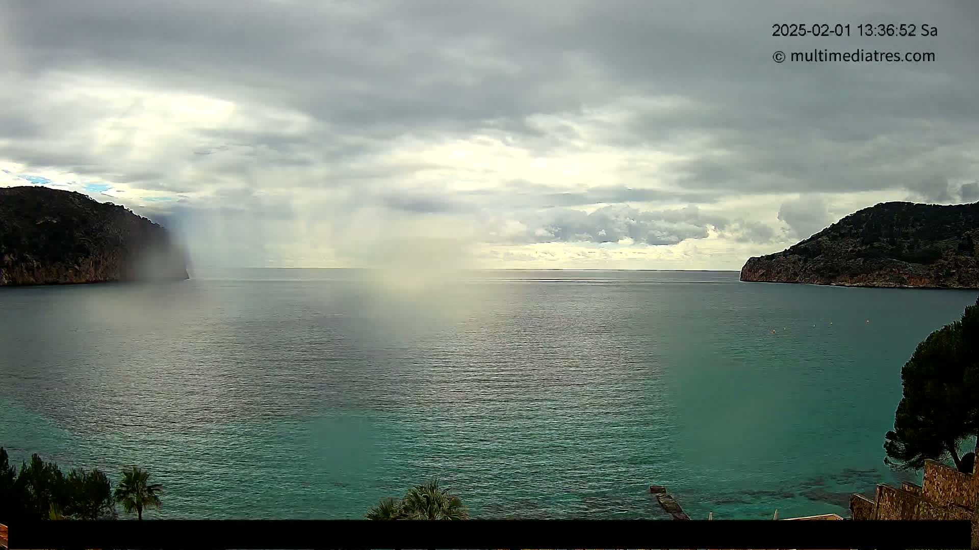 A body of teal water is seen between two rocky hills under a cloudy sky, with light rain or mist falling near one of the hills.