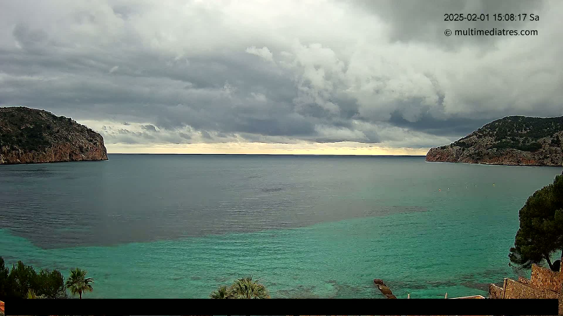 A calm bay with teal water is nestled between two rocky hills under a cloudy sky.