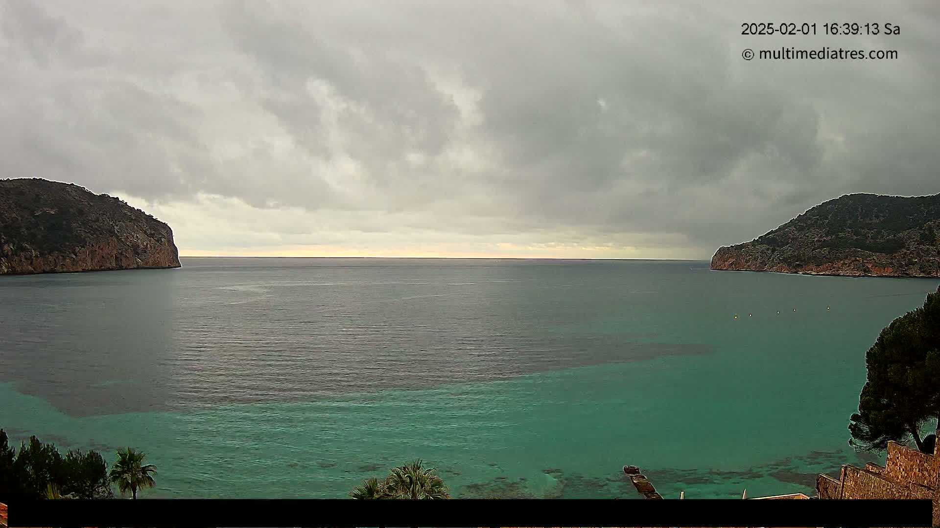 A tranquil bay with teal-colored water is nestled between two rocky hills under an overcast sky.