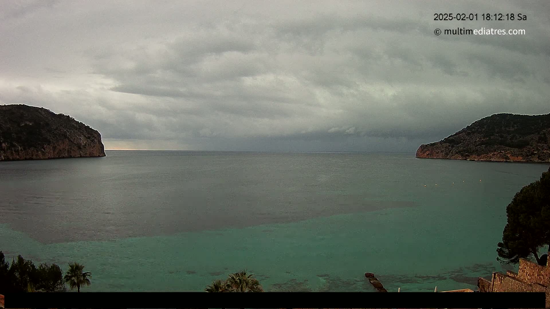 A calm sea with varying shades of green and gray is nestled between two dark, rocky hills under a cloudy sky.