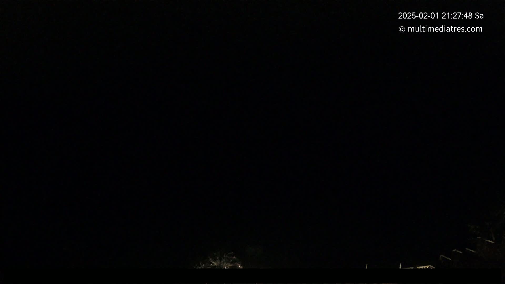 A dark nighttime scene shows a few dimly lit structures at the bottom of the frame against an entirely black sky.