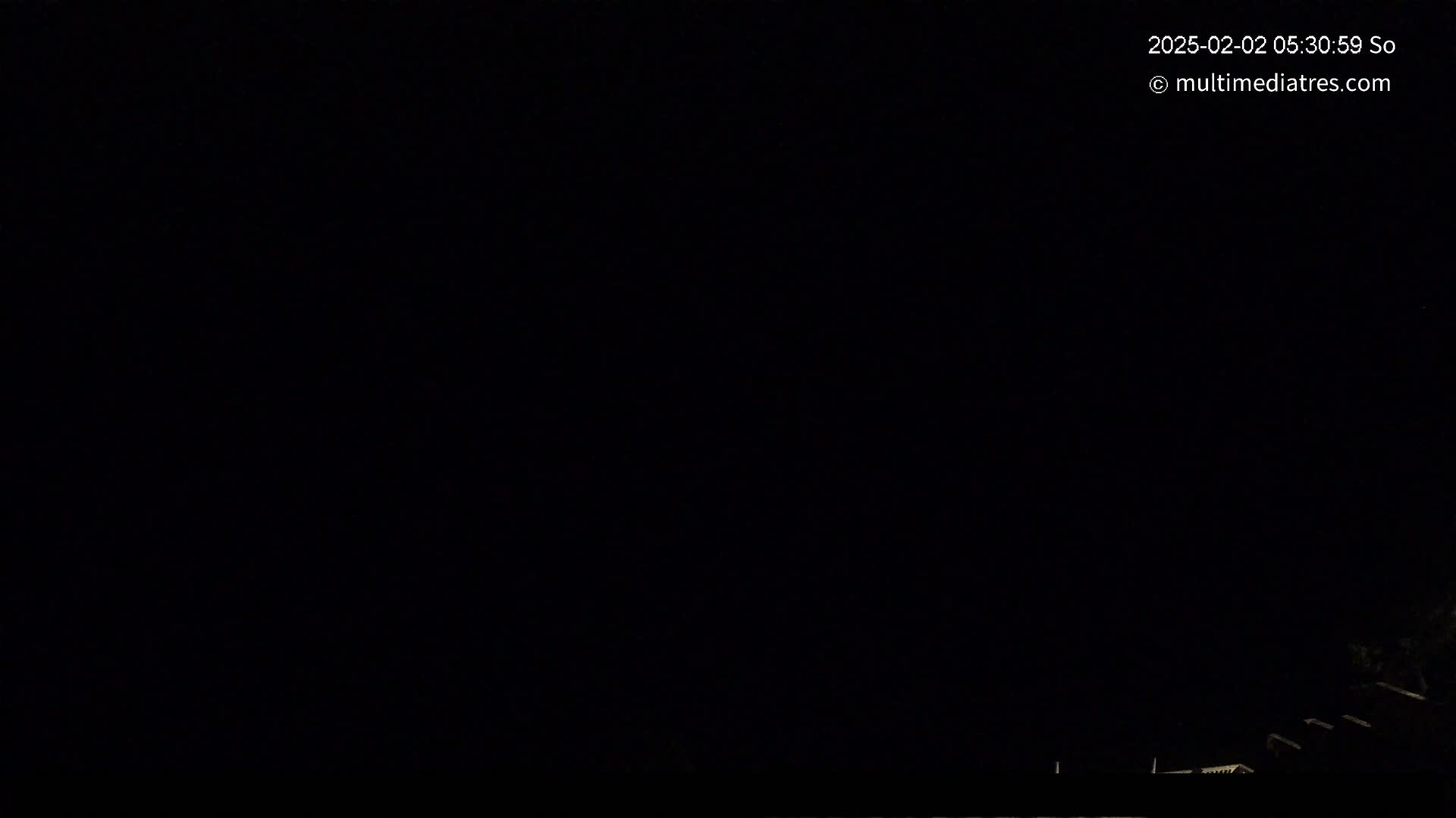 A dark nighttime scene shows the silhouettes of trees and rooftops at the very bottom of the frame against a completely black sky.