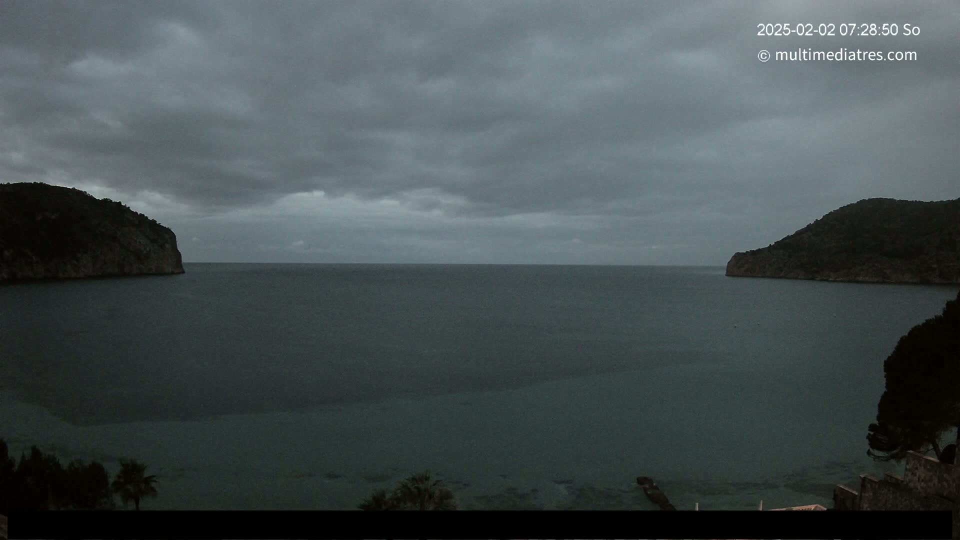 A dark, overcast sky hangs over a calm bay nestled between two dark, rocky hills.