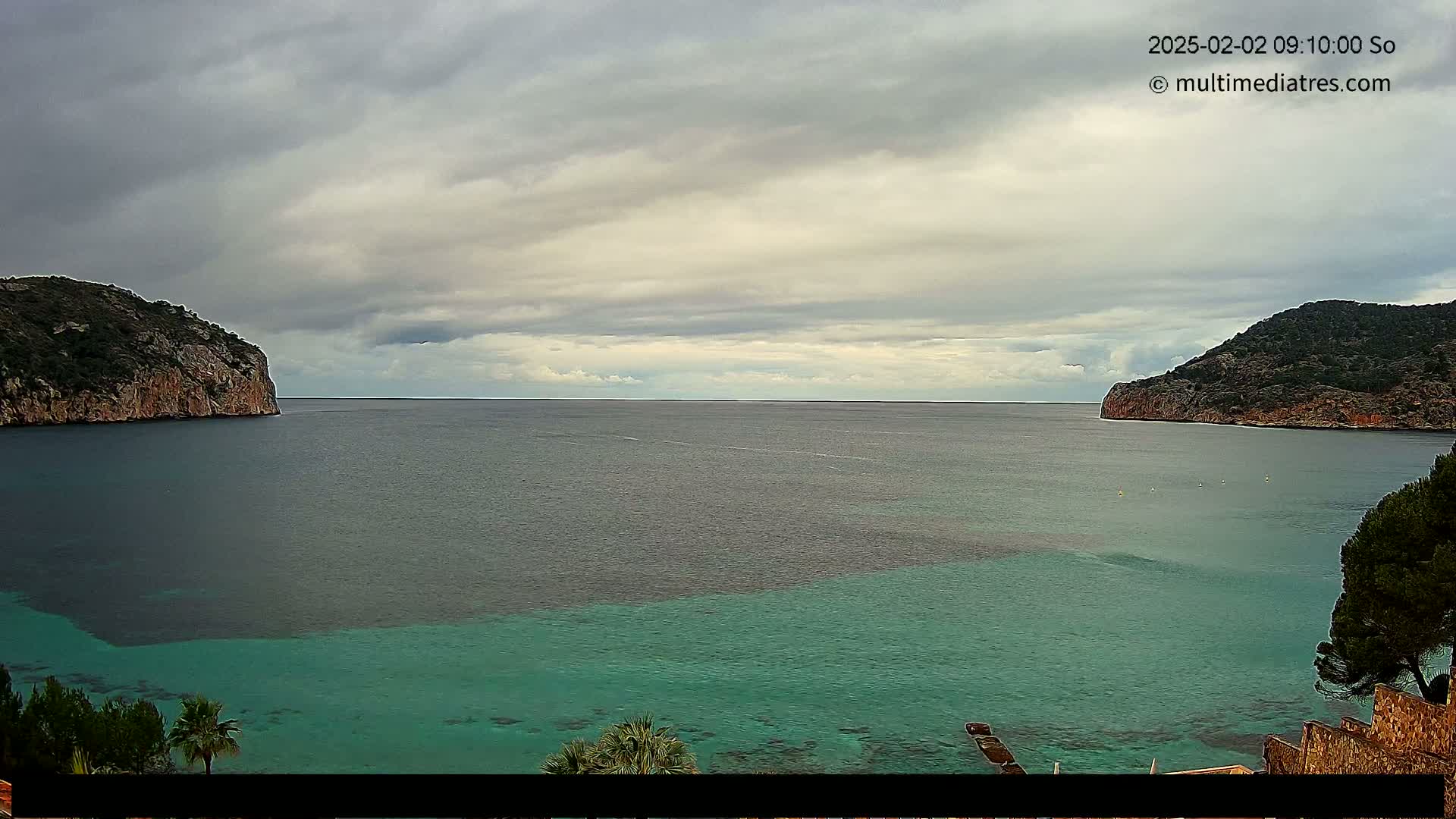 A calm bay with teal water is nestled between two rocky hills under a cloudy sky.