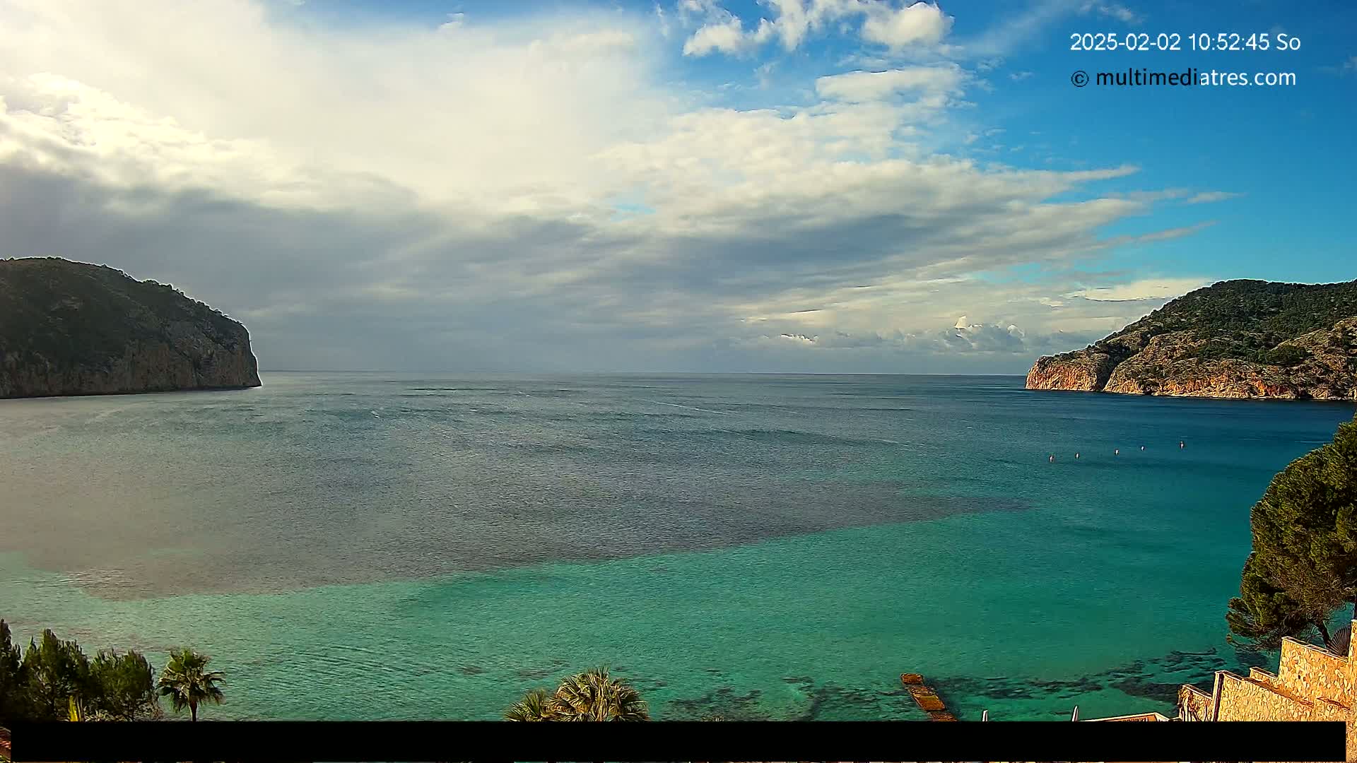 A partly cloudy day shows a tranquil bay of teal water between two rocky hillsides, with a few people visible in the distance.