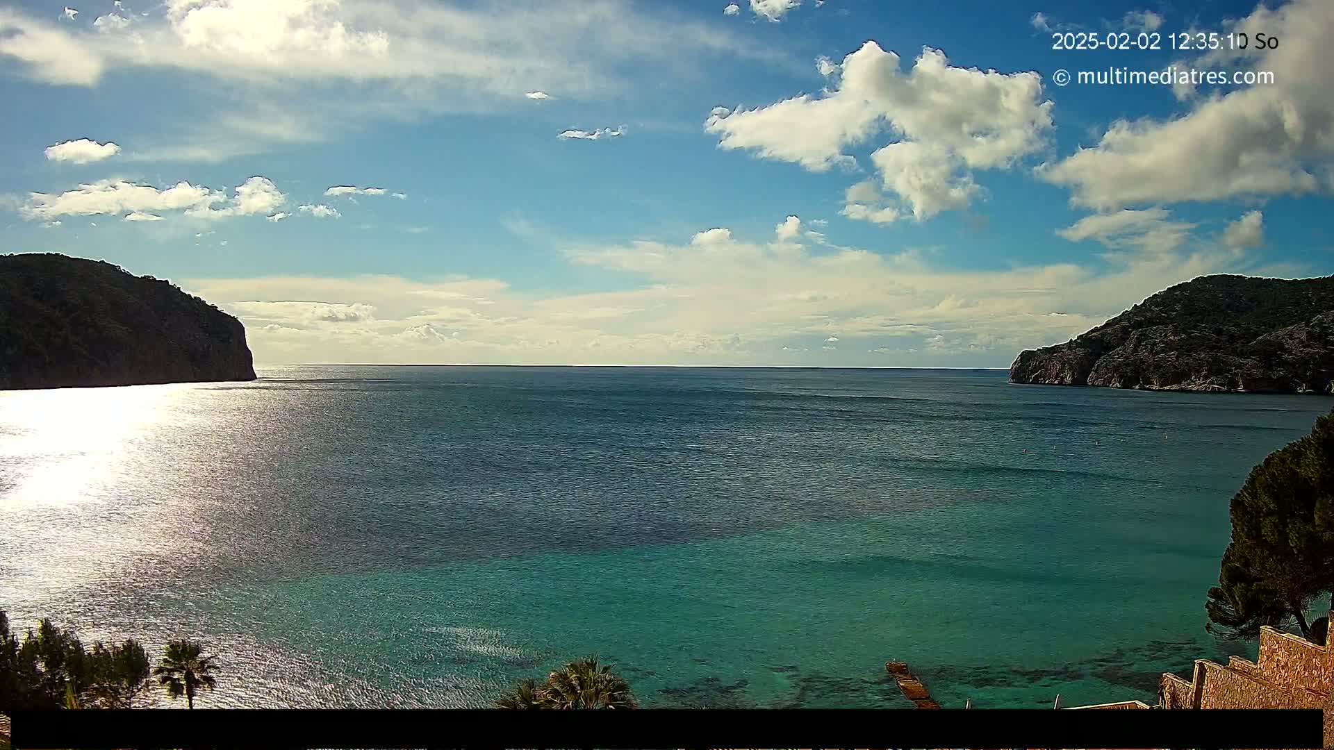 A partly cloudy sunny day reveals a tranquil bay of teal water nestled between two dark green cliffs.