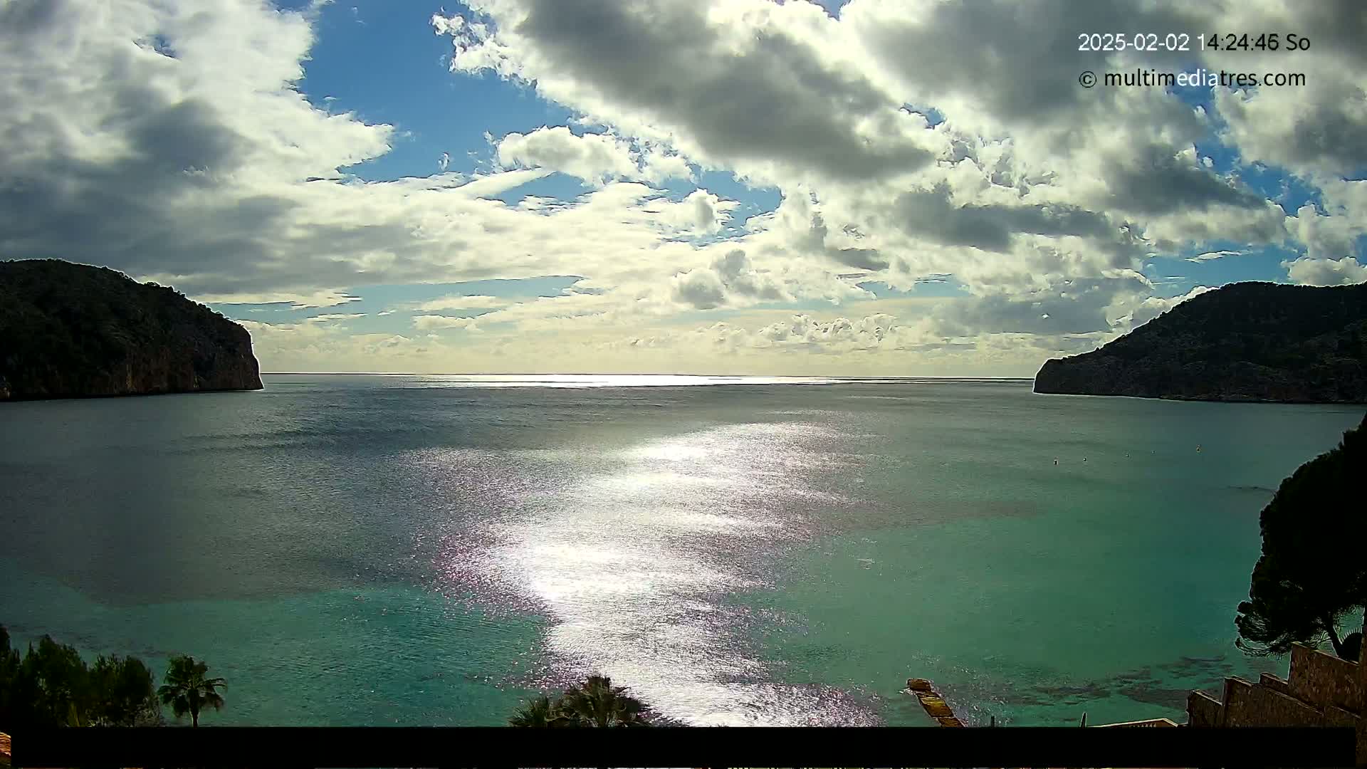 A partly cloudy day shows a tranquil bay of teal water reflecting sunlight, enclosed by dark green hills.
