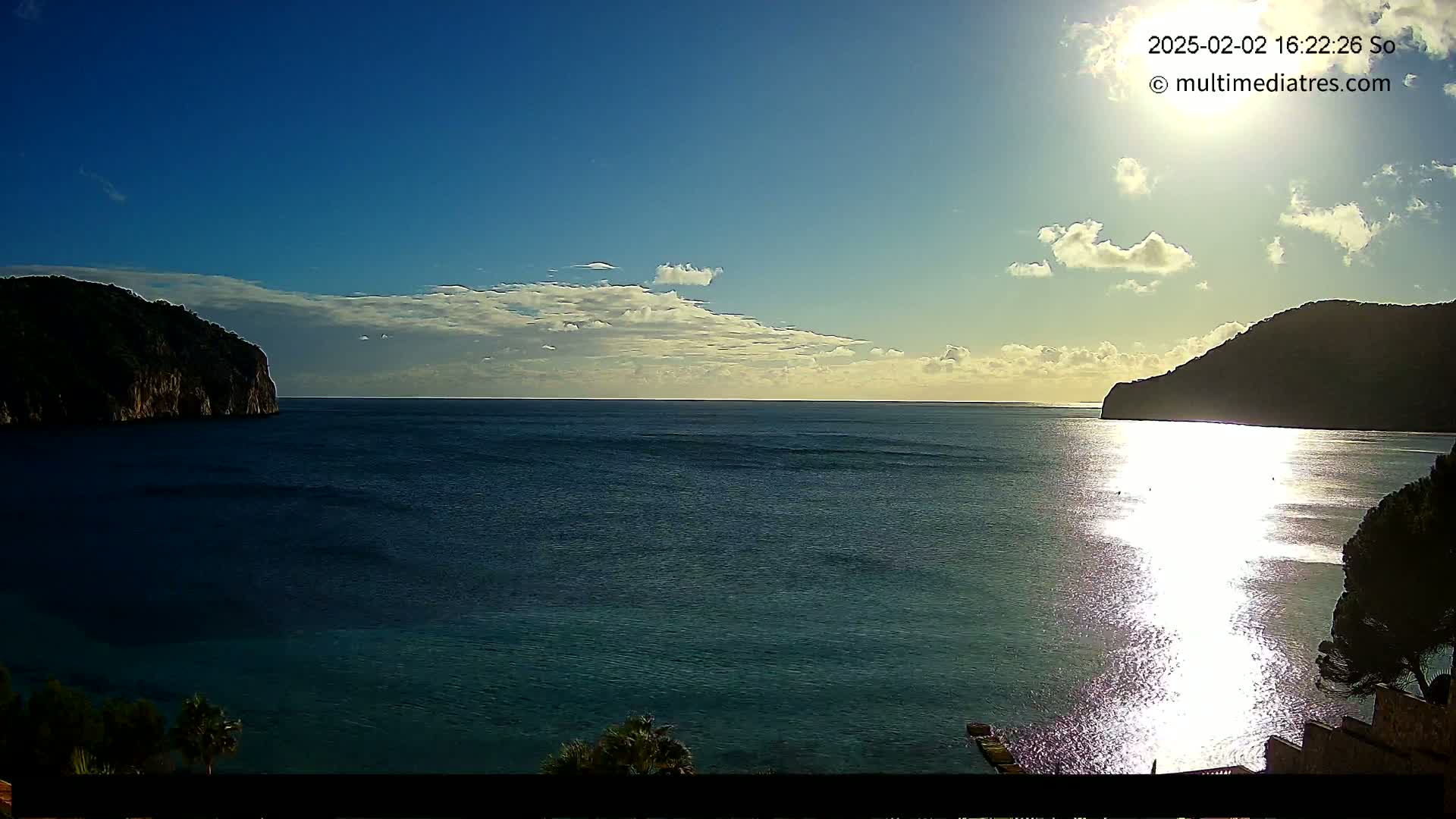 A calm sea between two dark, rocky hills under a partly cloudy sky with the sun shining brightly on the water.