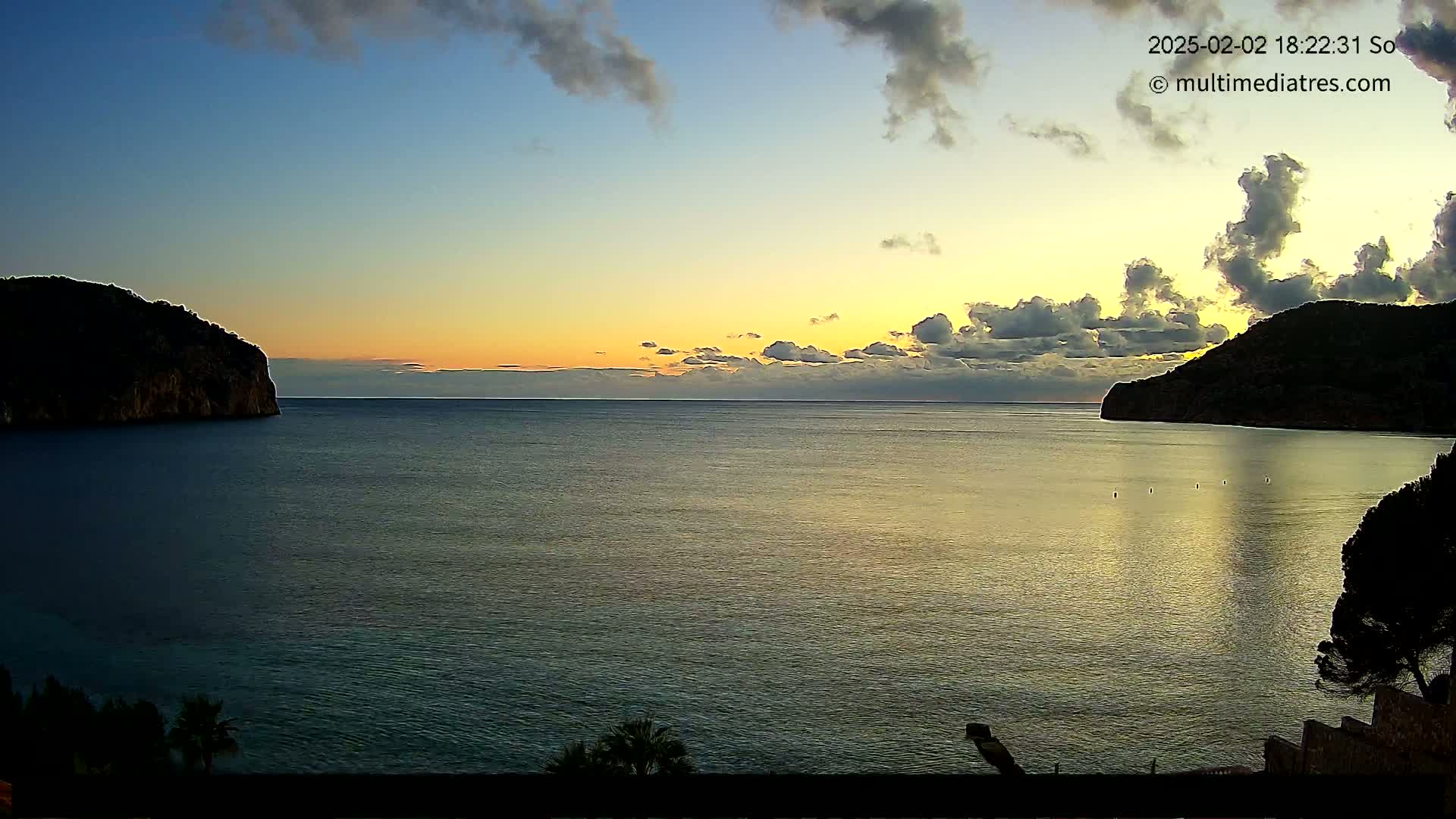 A calm sea at sunset, nestled between two dark, rocky hills under a partly cloudy sky.