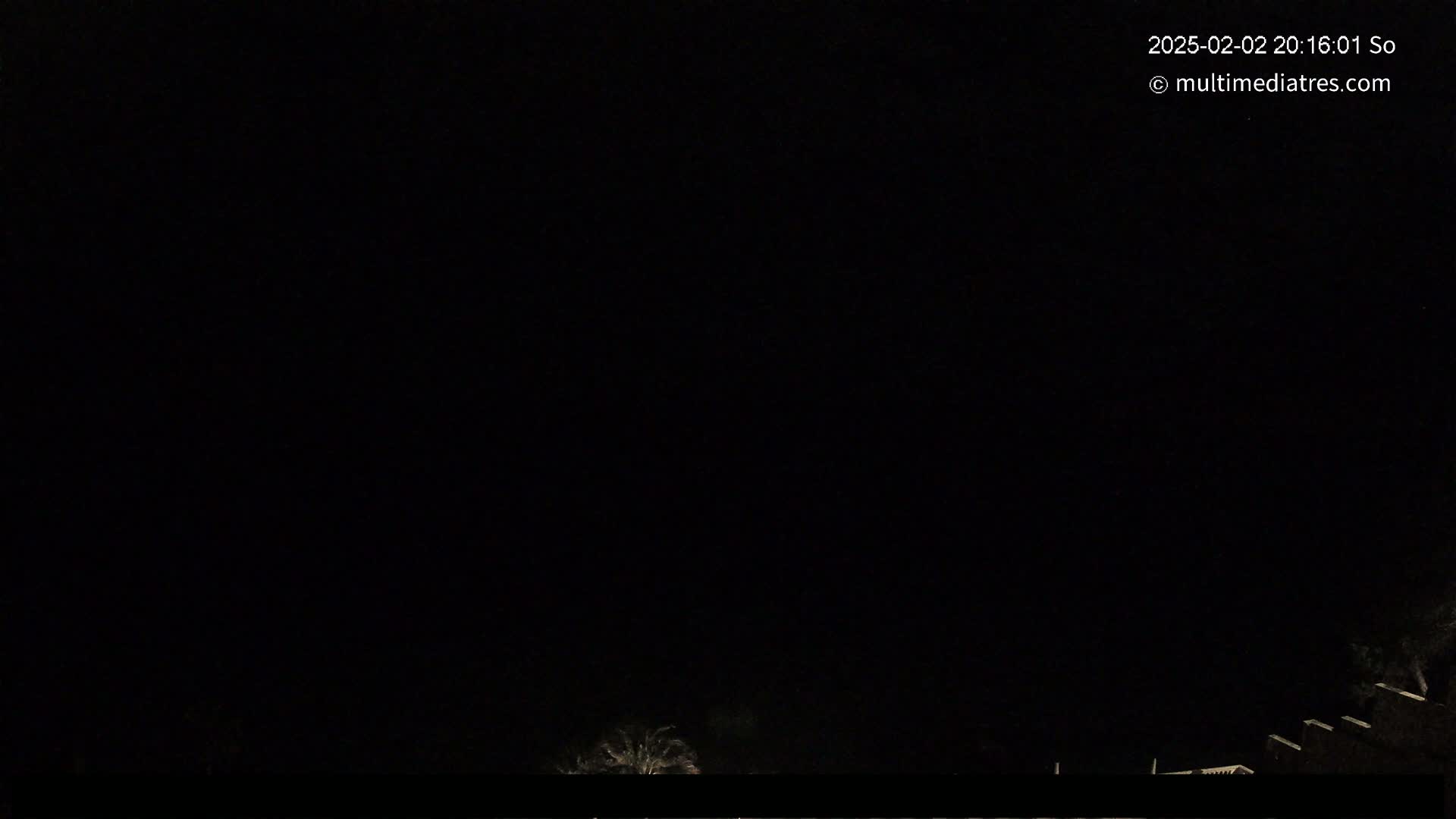 A nighttime outdoor scene shows a mostly dark sky with faintly visible trees and buildings at the bottom.