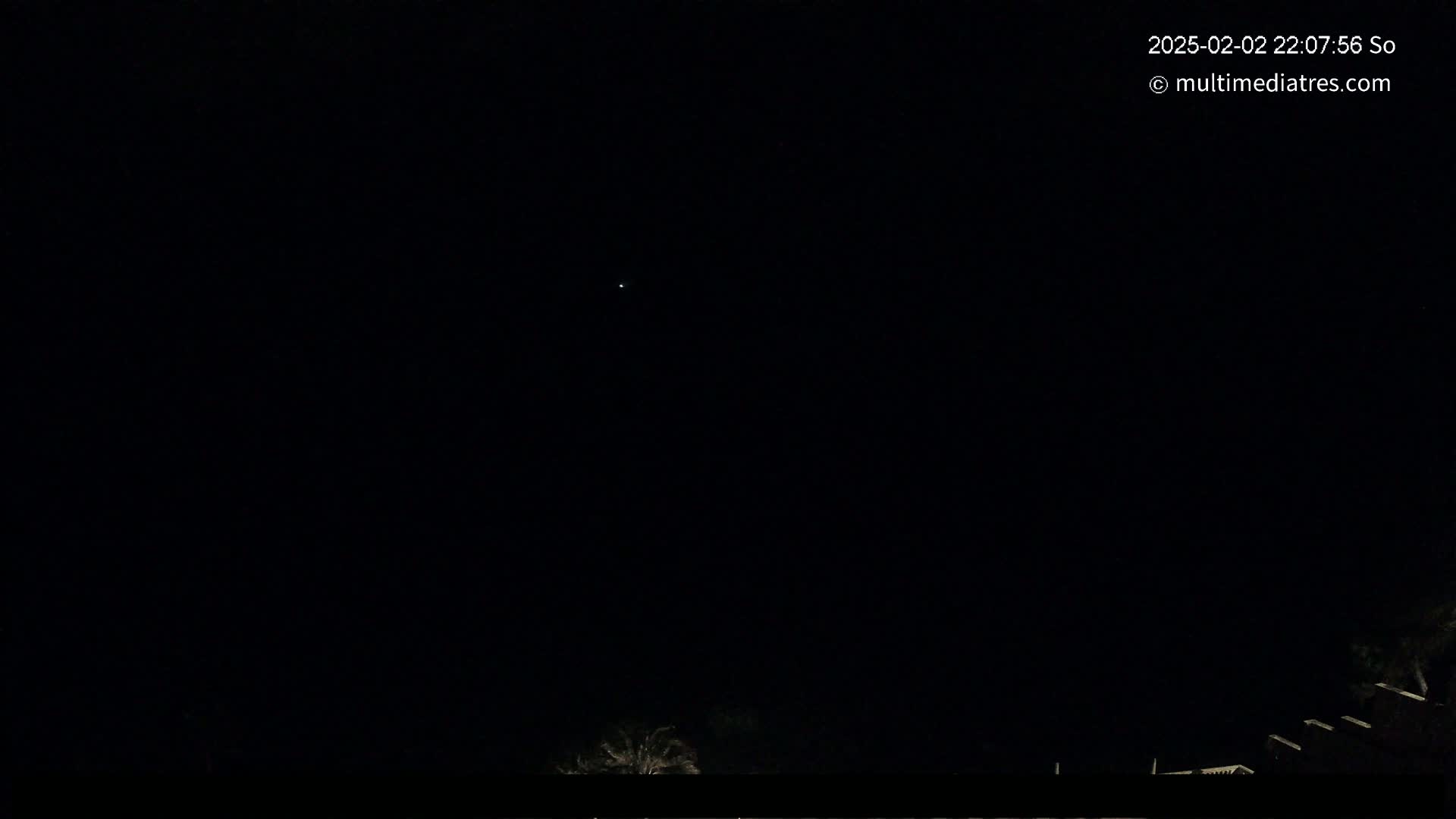 A dark night sky shows a single bright star above a low horizon with some dark silhouettes of trees and buildings.