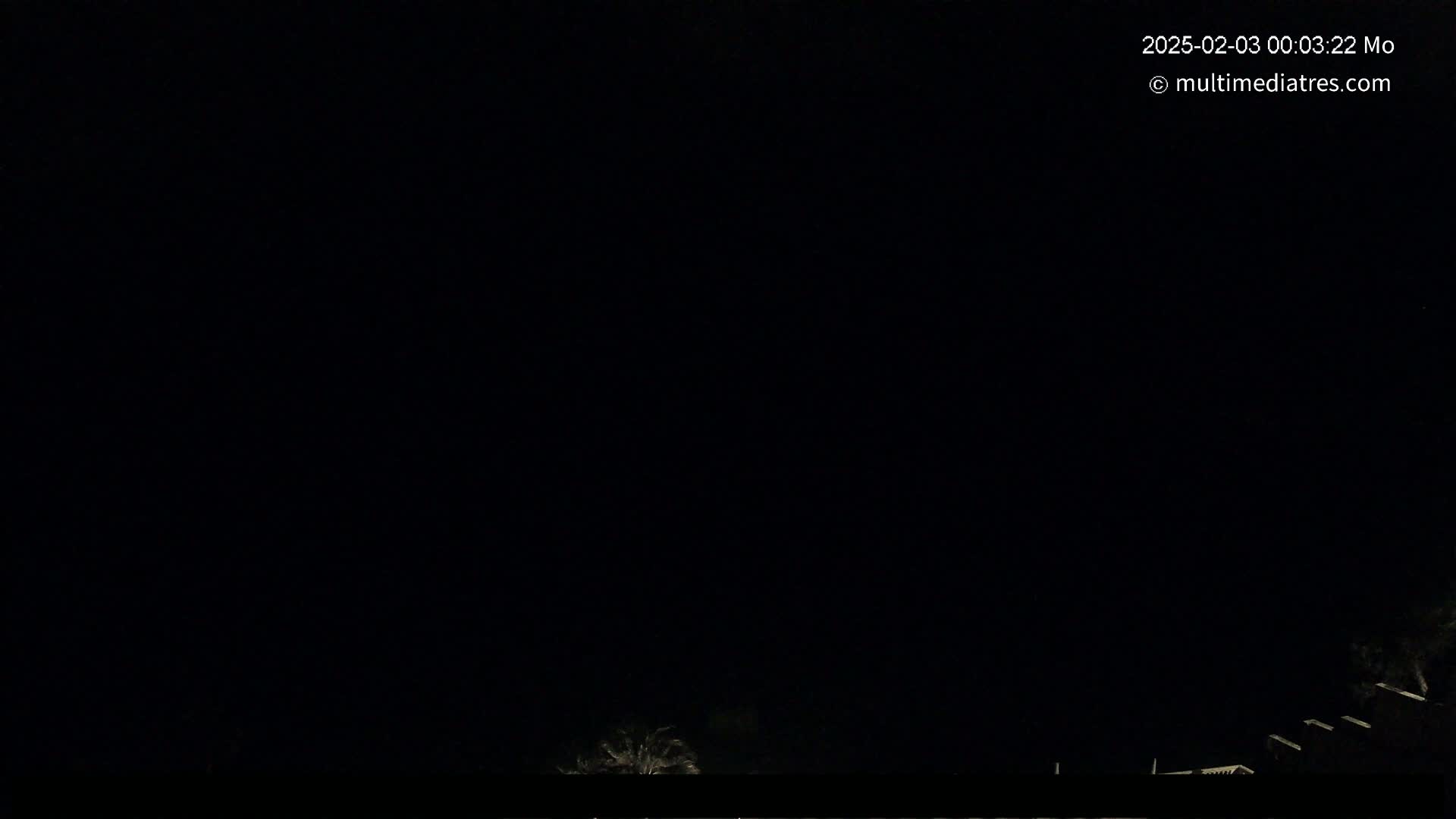 A nighttime outdoor scene shows a mostly dark sky with a few dimly lit trees and buildings visible at the bottom of the frame.