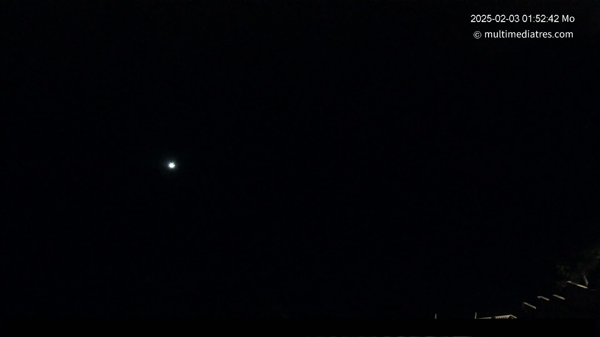 A bright light, possibly a star or planet, is visible in a dark night sky above a dark silhouette of rooftops.