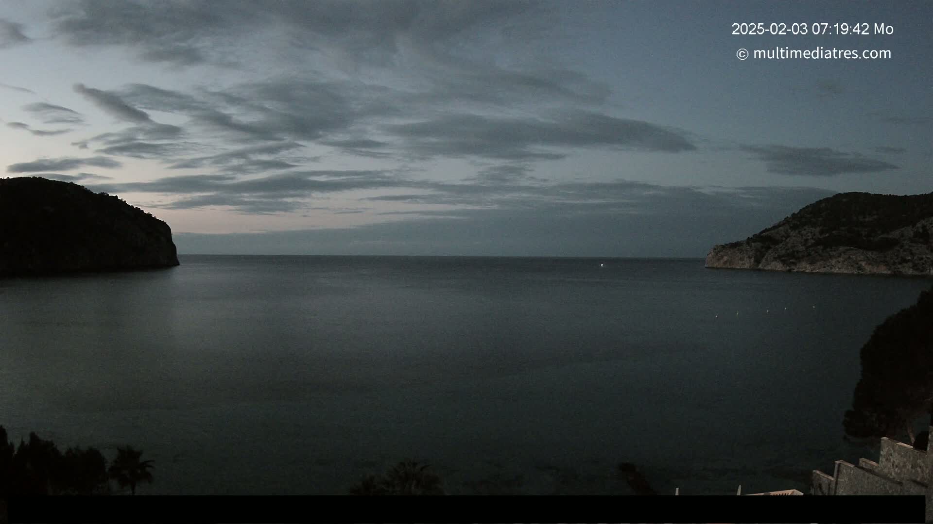 A dark, calm bay is nestled between two mountainous silhouettes under a cloudy, twilight sky.