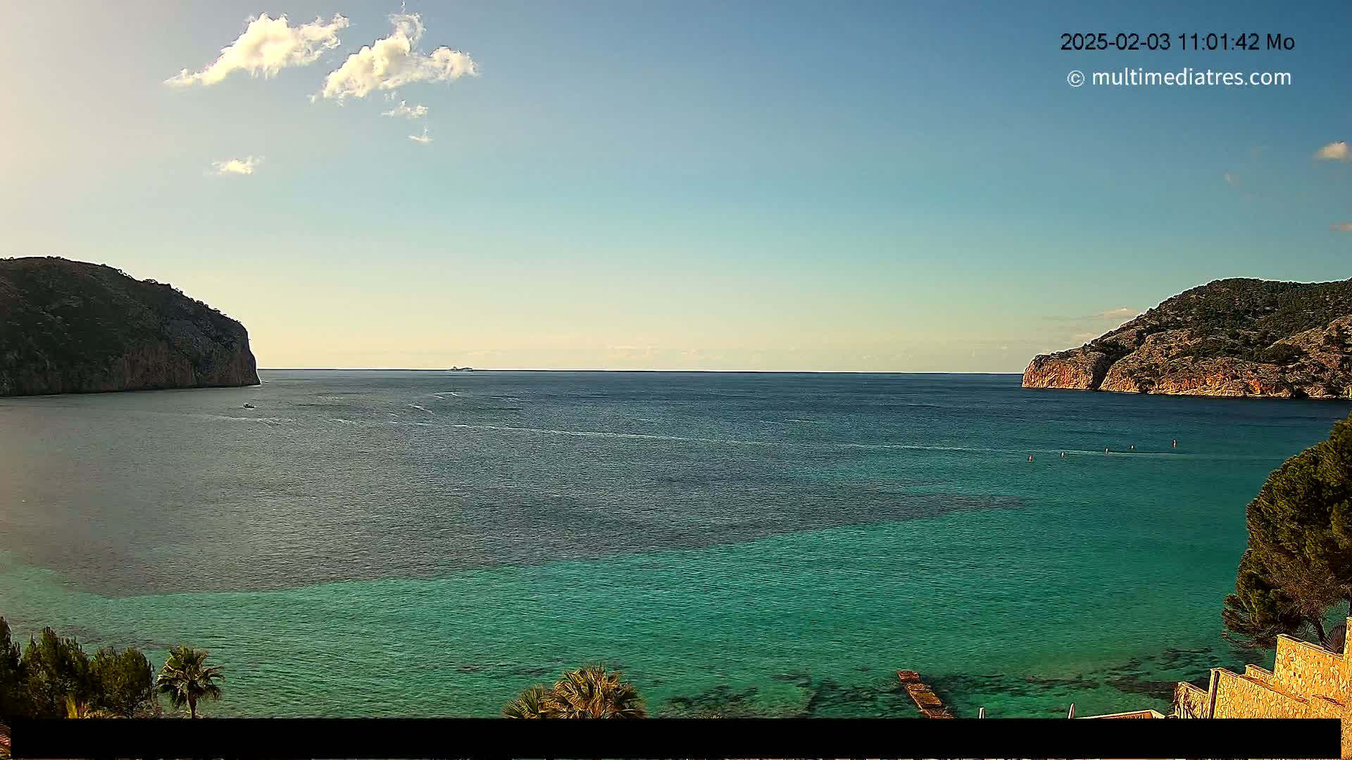 A calm, partly cloudy day shows a tranquil bay of teal and dark blue water between two rocky hillsides.