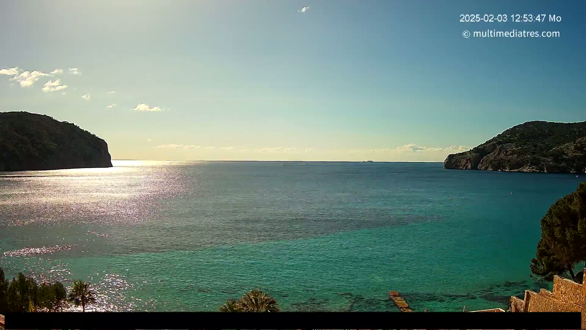 A tranquil, sunny day reveals a turquoise bay nestled between two rocky hills, with a few boats visible in the distance.