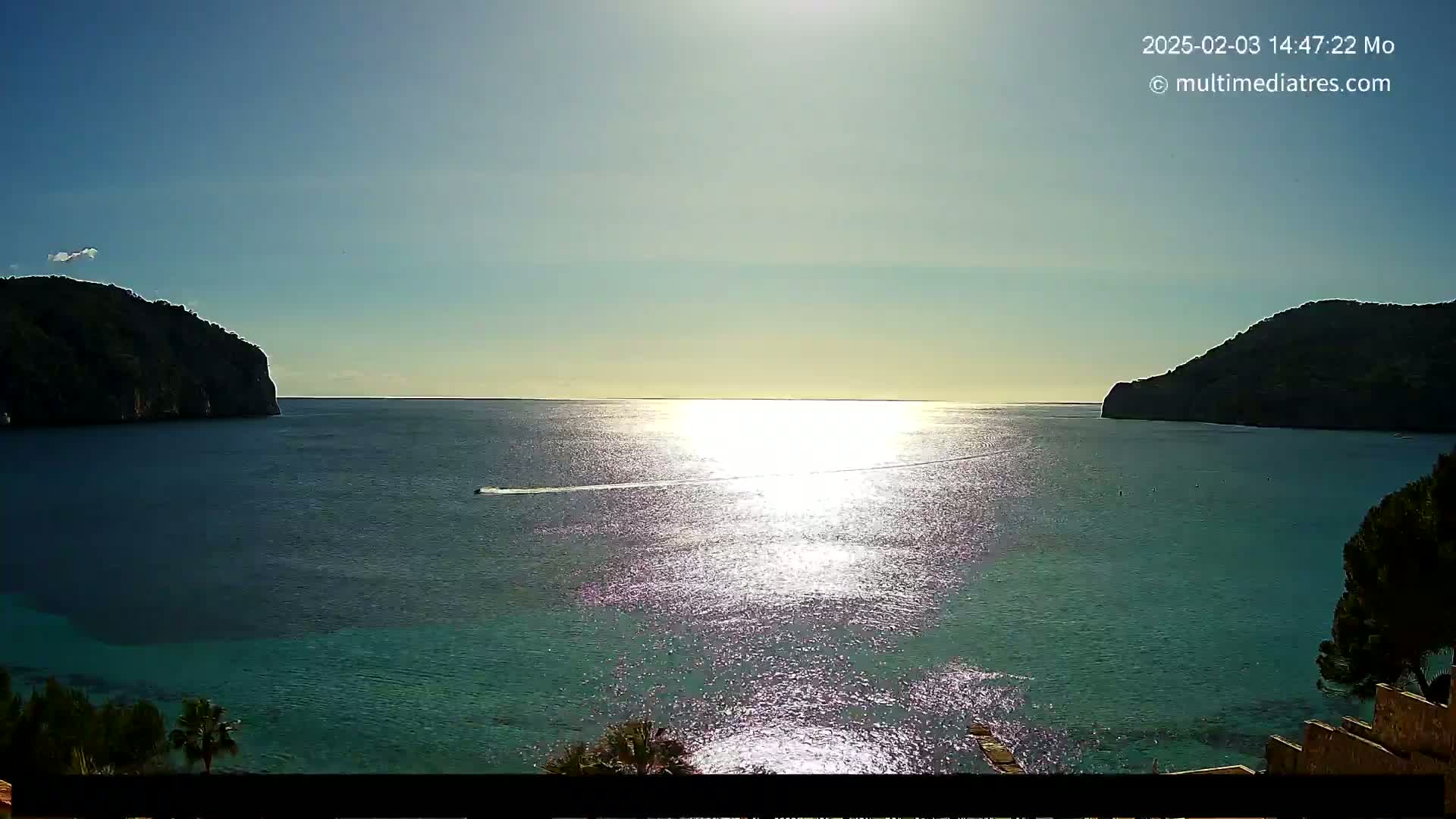 A sunny day reveals a tranquil bay with calm, sparkling water between two dark, verdant hills, and a small boat leaves a wake in the distance.